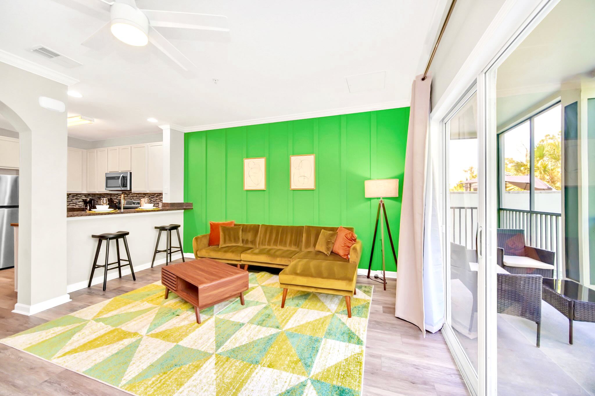 A bright open-concept living area features a chartreuse velvet sectional sofa, a mid-century modern coffee table, and a vibrant geometric rug, with a lime green accent wall. The space flows into a kitchen with a breakfast bar and stainless steel appliances.