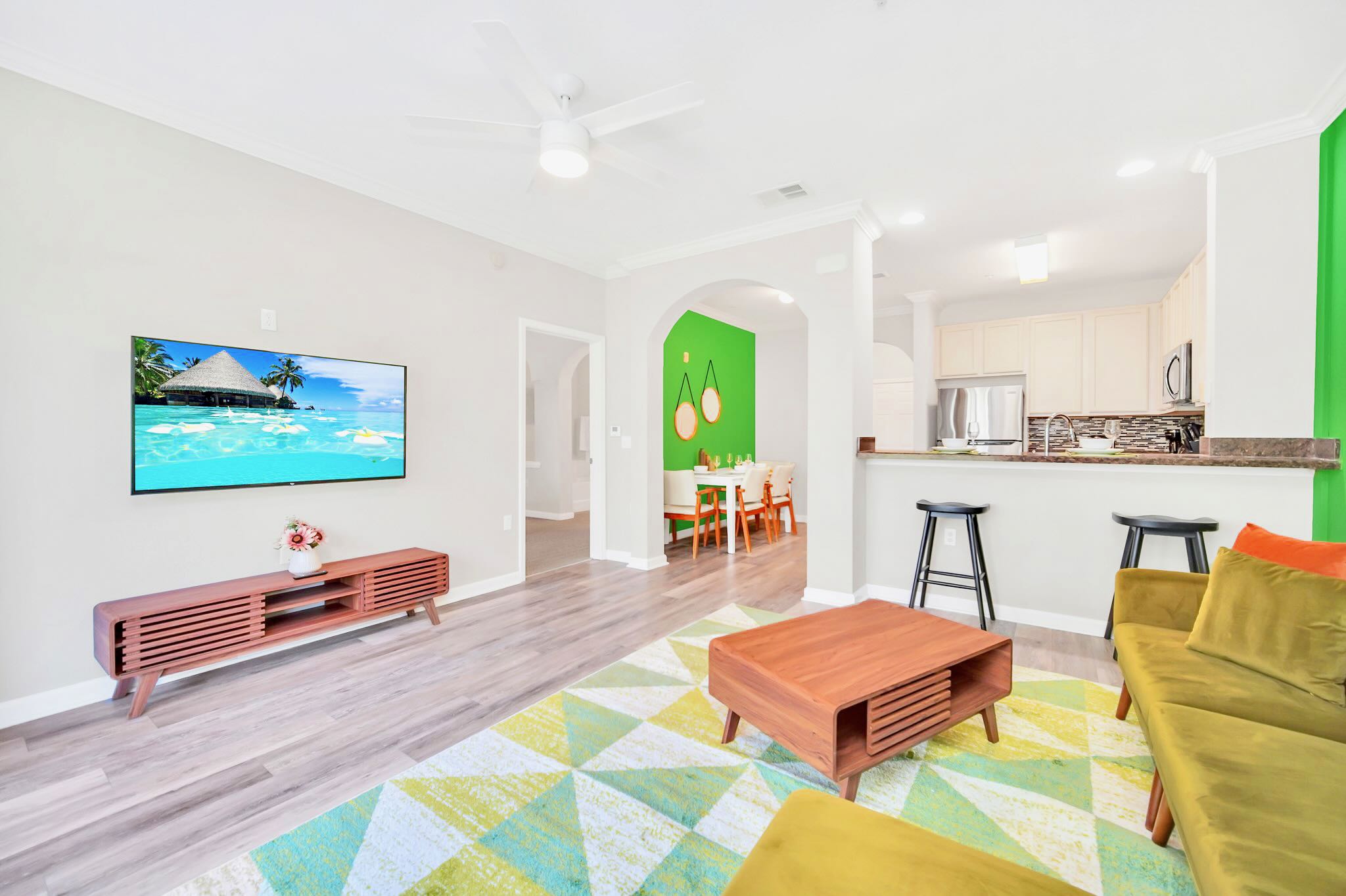 An expansive open-concept living space features a chartreuse sectional sofa and a geometric rug, flowing into a dining area with a lime green accent wall and a modern kitchen with a breakfast bar. Light wood-look flooring unifies the entire area.