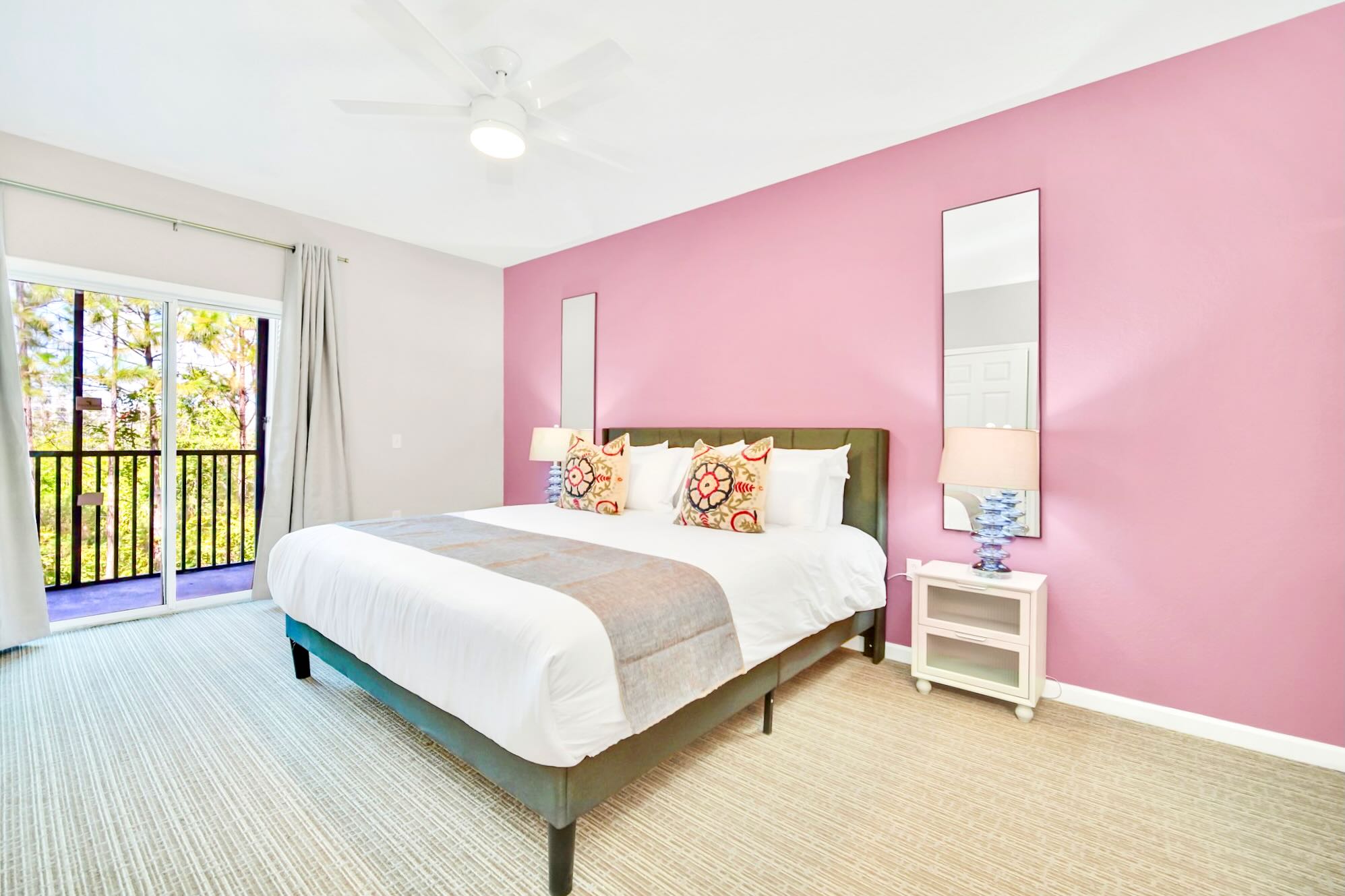 Master bedroom with a king-sized bed, a vibrant pink accent wall, two tall mirrors, and a balcony access, featuring light carpet and two bedside tables with lamps.