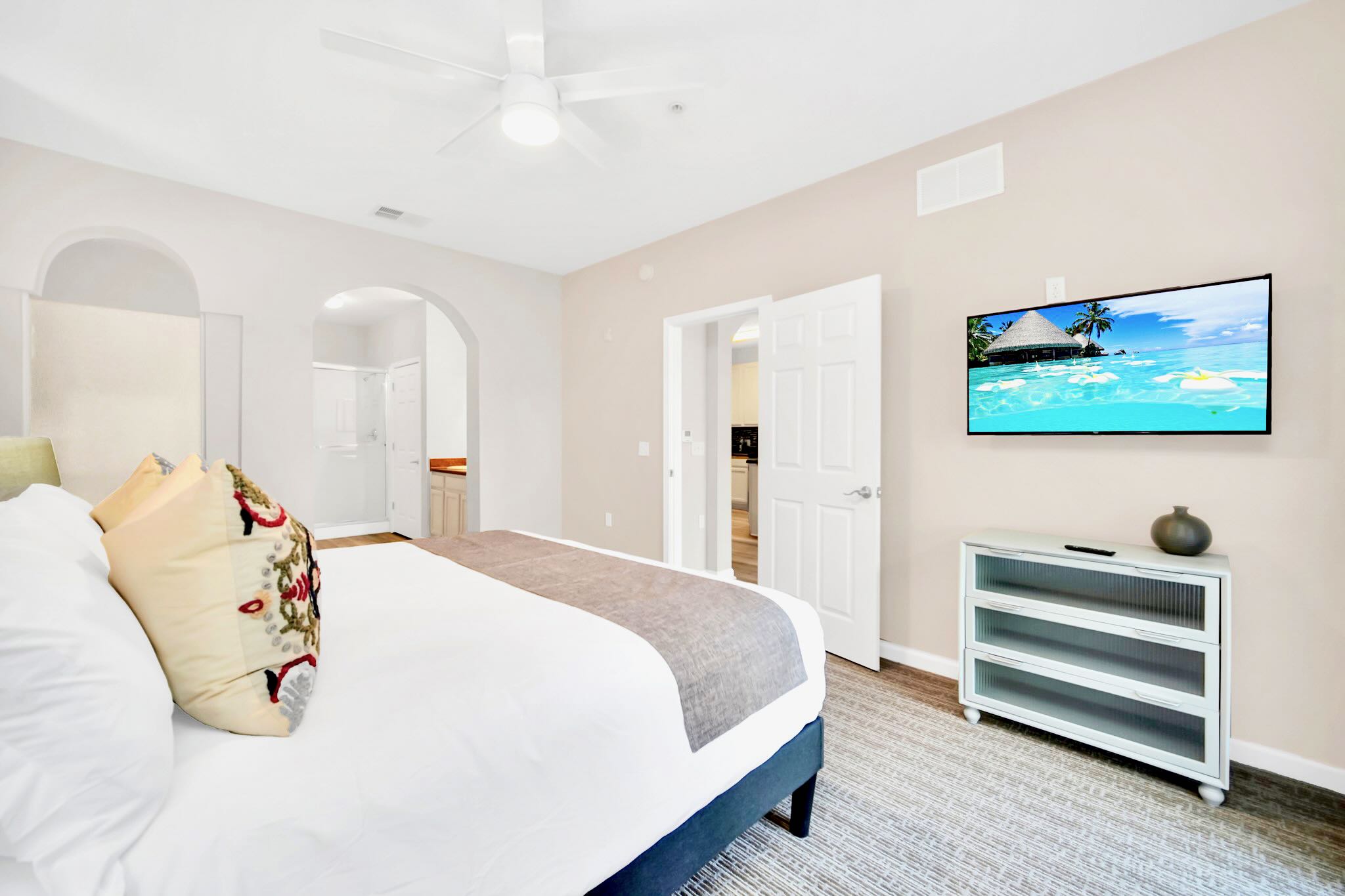 Master bedroom with a large bed, light gray carpet, and a wall-mounted TV displaying a beach scene, with an arched doorway leading to the en-suite bathroom.
