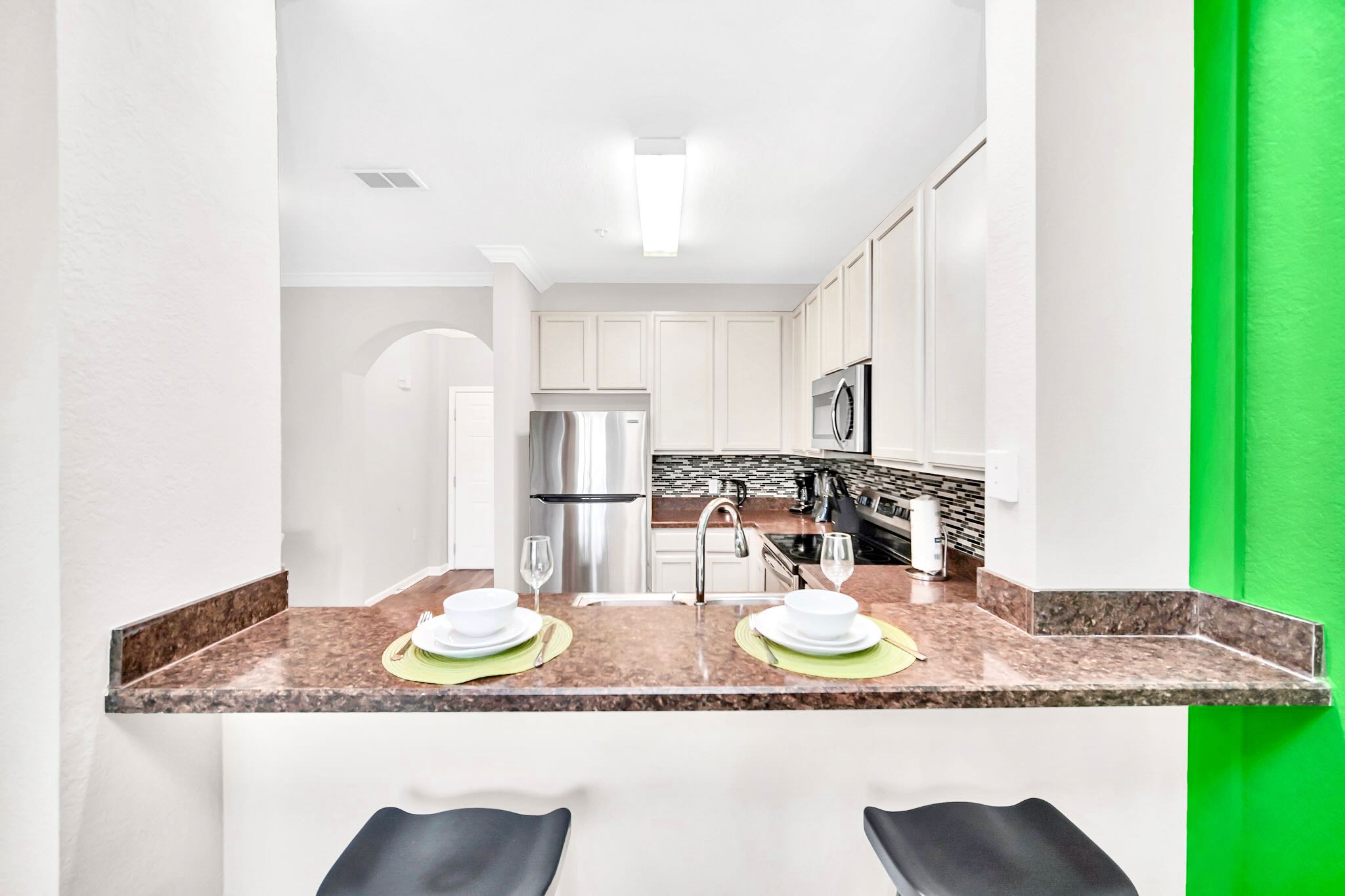 View of the kitchen from a breakfast bar, showing white cabinets, stainless steel appliances, and dark granite countertops, with two place settings on the bar.