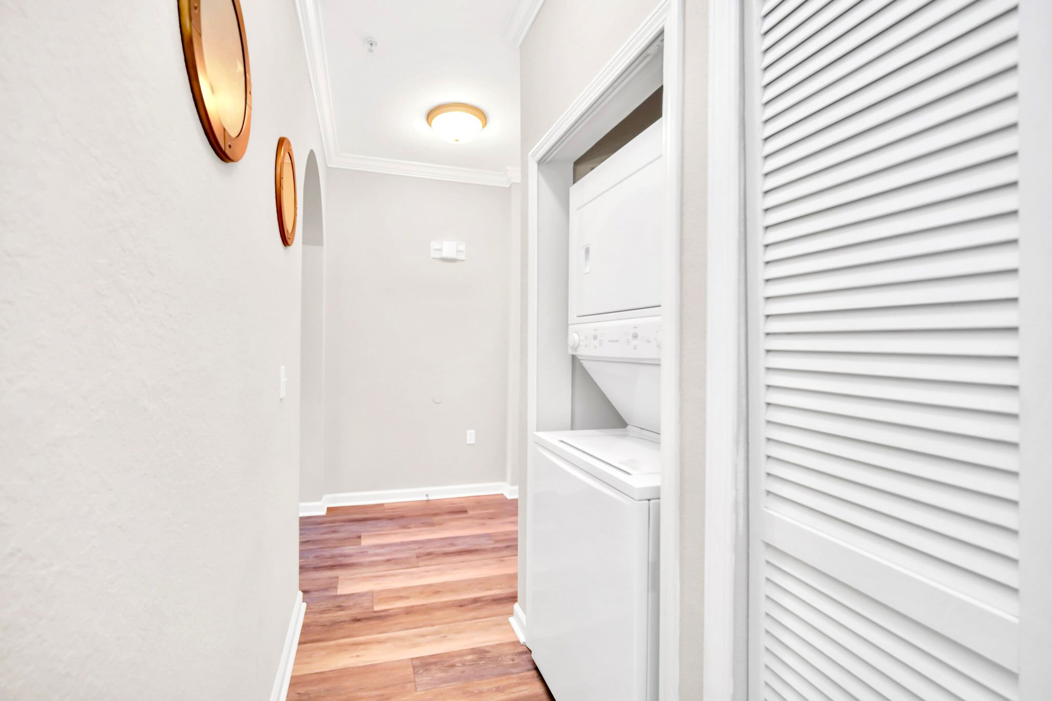 A narrow hallway with light gray walls and light wood-look flooring leads to a closet containing a stacked white washer and dryer unit. Two round, gold-framed mirrors adorn the left wall, and a flush-mount ceiling light illuminates the space.