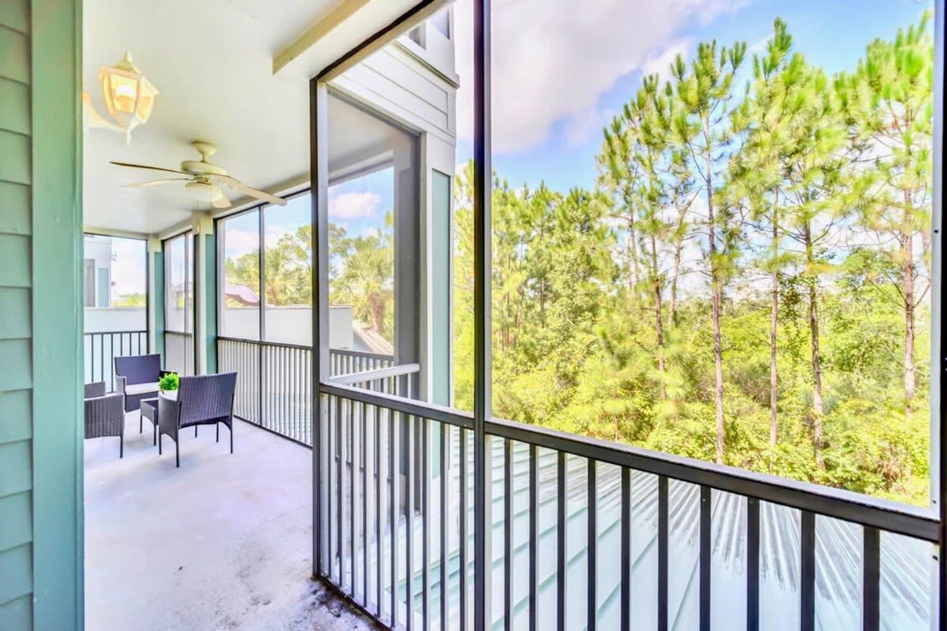 A screened balcony provides an inviting outdoor space with a ceiling fan and seating, offering expansive views of dense green trees and the surrounding natural landscape. The black metal railing ensures safety while maintaining an open feel.