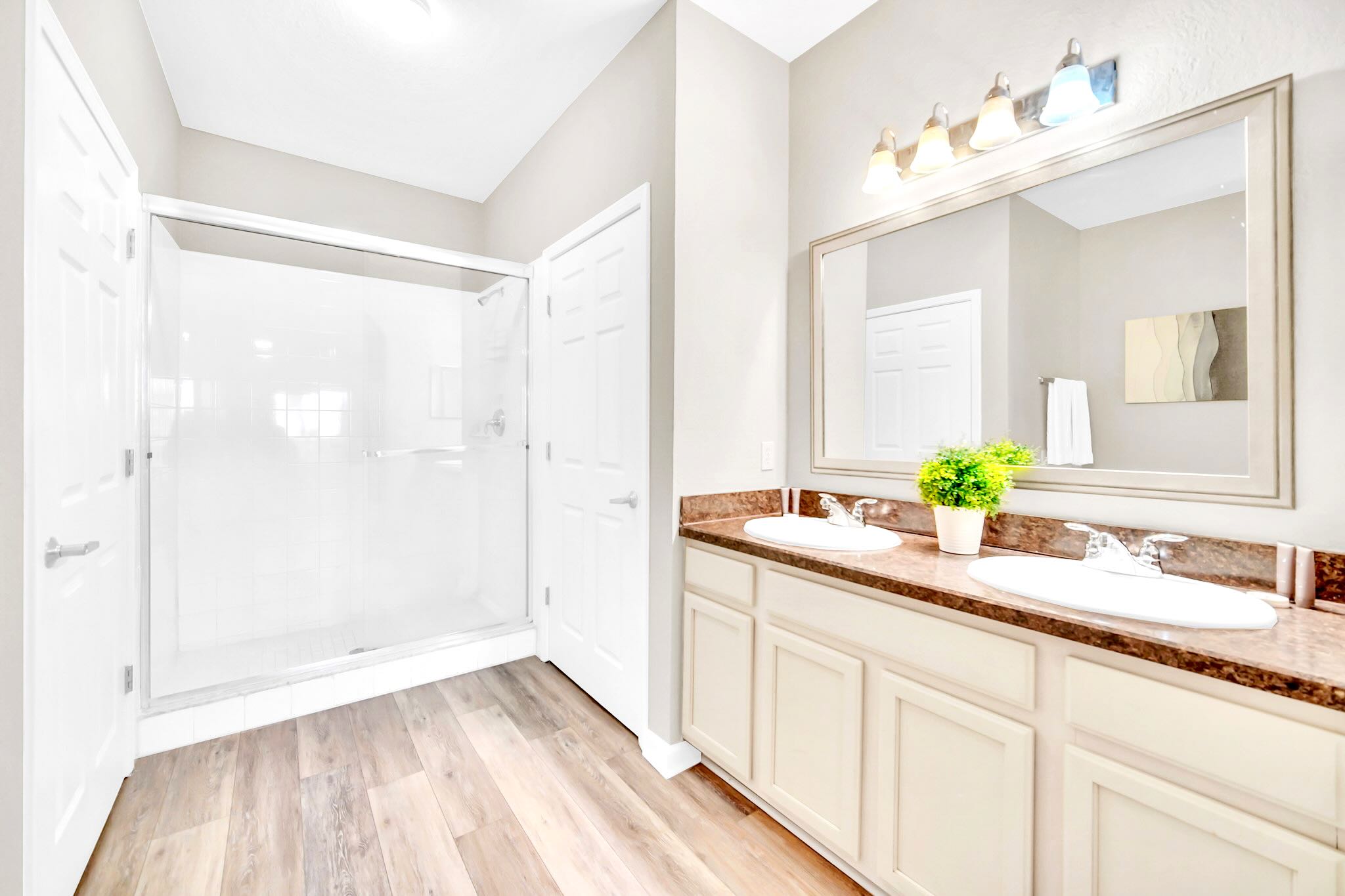 Spacious en-suite bathroom with a double vanity, large mirror, and a walk-in shower with a glass door, featuring light wood-look flooring and light-colored walls.