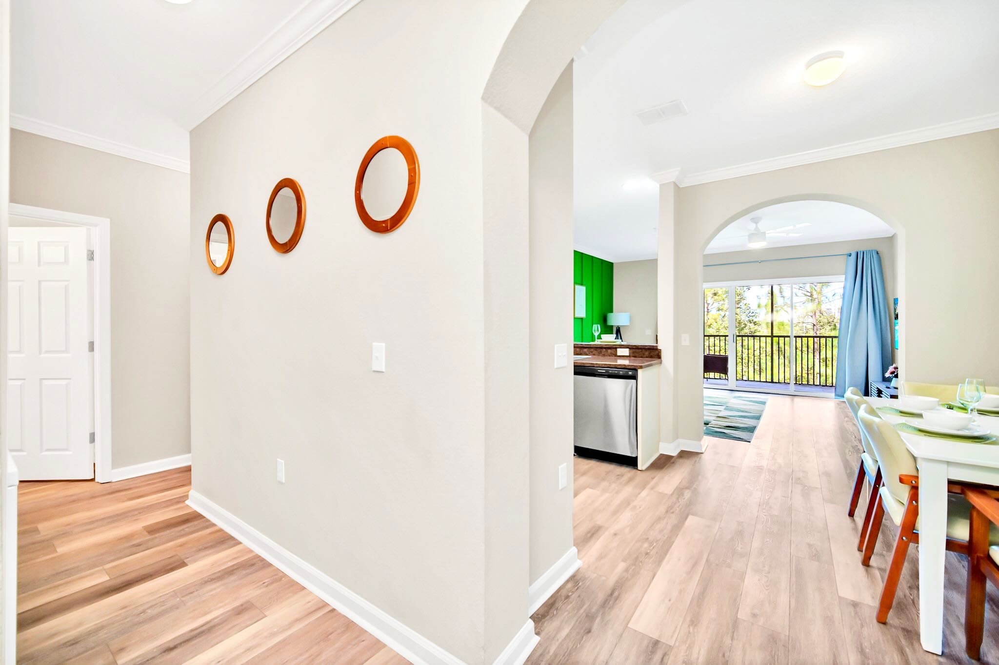 Bright hallway with light gray walls and light wood-look flooring, featuring three round wooden mirrors on the wall and an arched entryway leading to the kitchen and dining area.