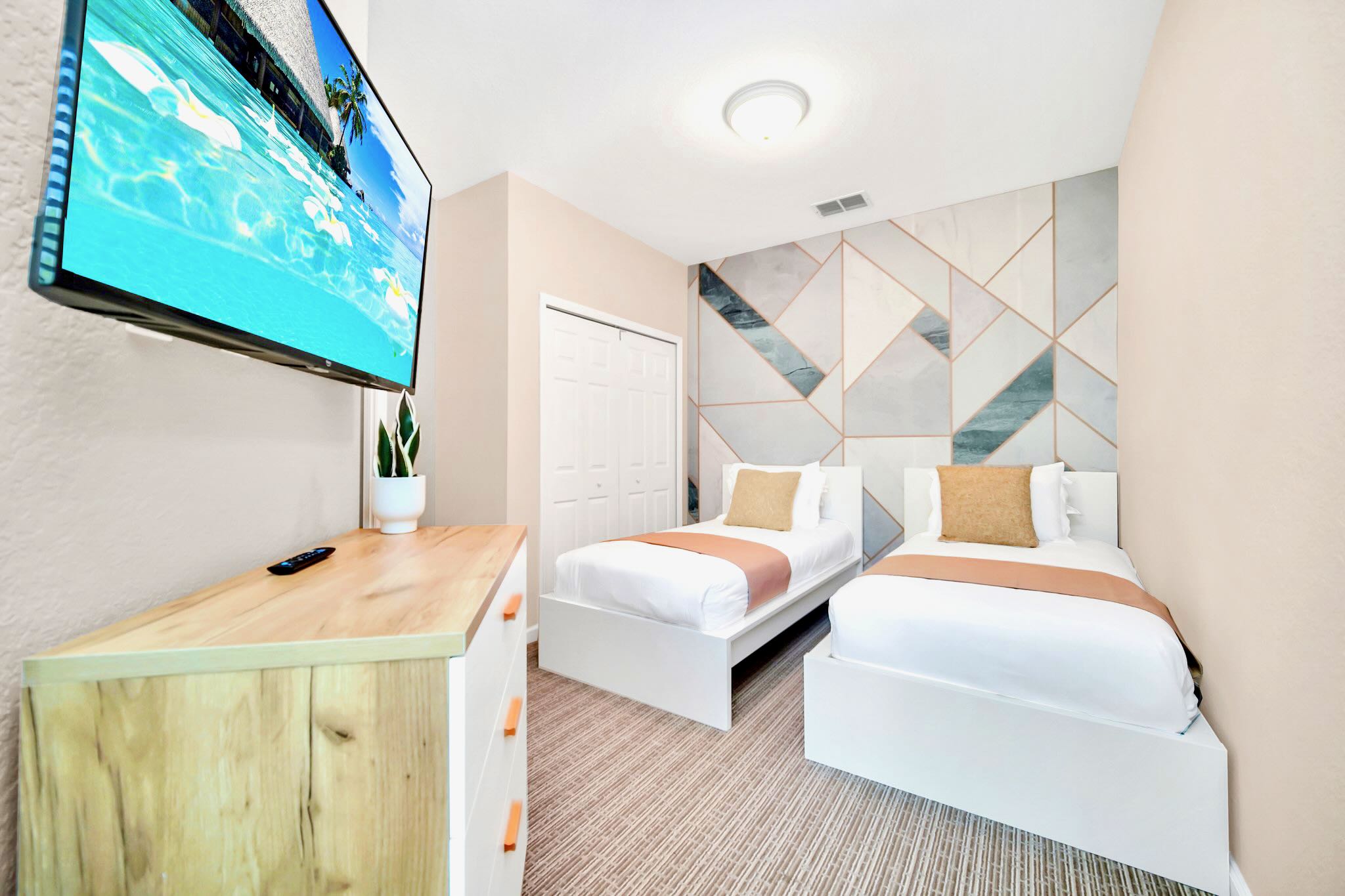 View of a bedroom with two twin beds, a geometric accent wall, and a wall-mounted TV, showing a closet with bi-fold doors and light carpet flooring.