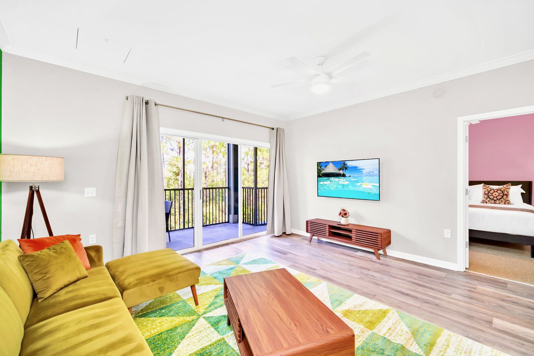 The living room showcases a chartreuse velvet sectional sofa and a geometric rug, with a flat-screen TV on a wooden console. A sliding glass door leads to a balcony, and an open doorway reveals a glimpse into a pink-walled bedroom.