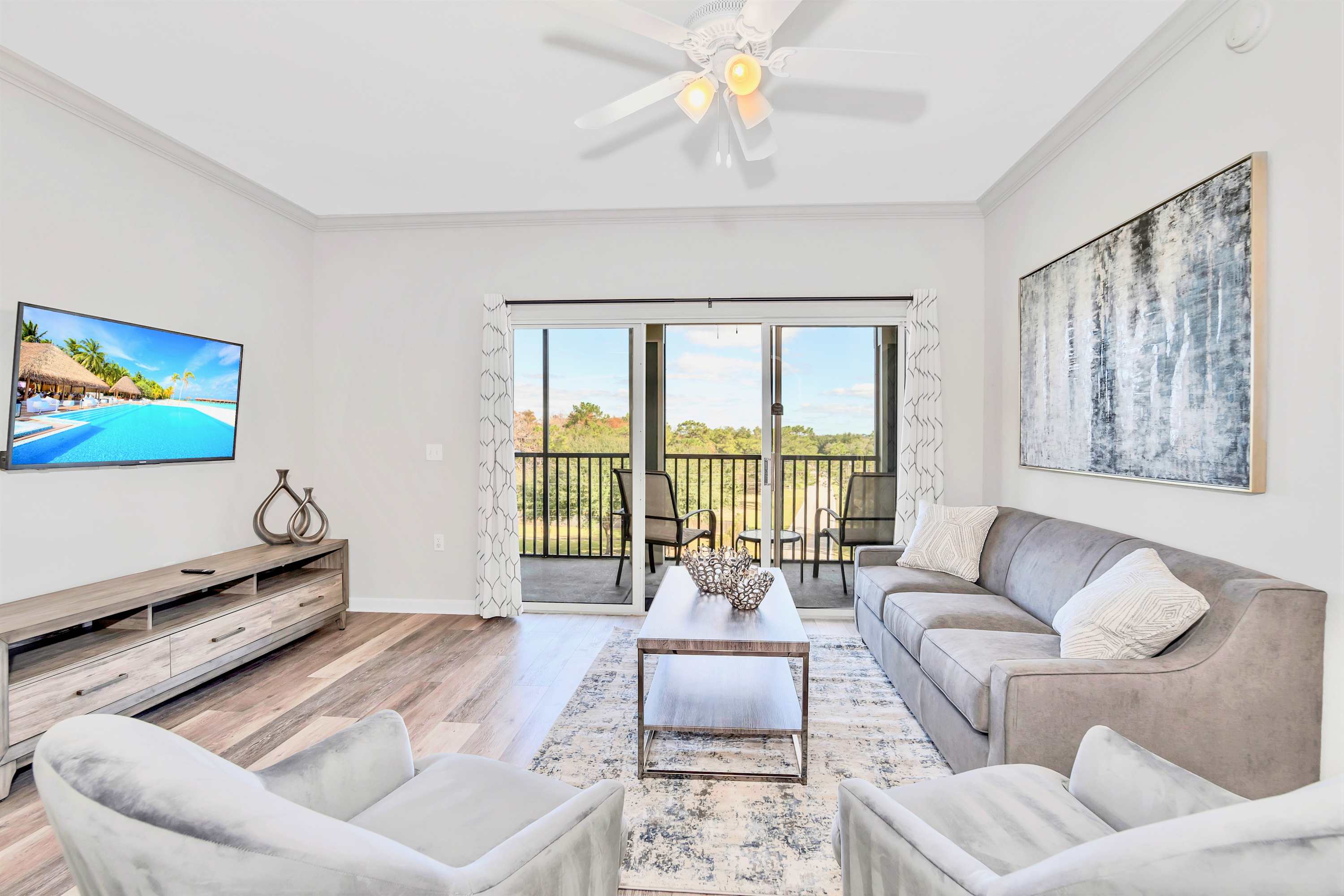 Bright living room featuring a gray sofa with throw pillows, two light gray armchairs, a wooden coffee table on a patterned rug, and a wall-mounted TV, with sliding glass doors opening to a balcony.