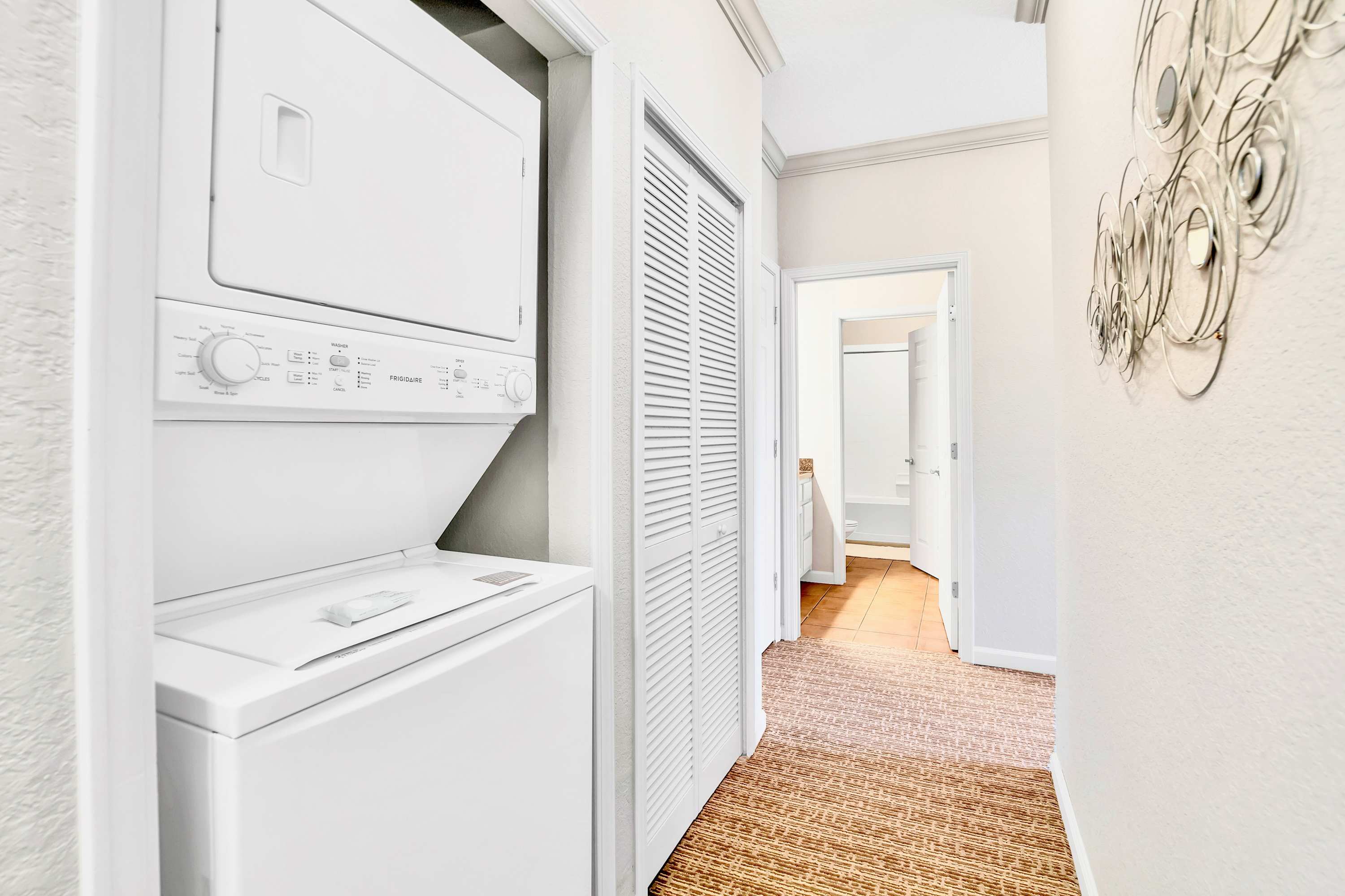 A bright hallway features a white stacked washer and dryer unit neatly tucked into a closet, with louvered bifold doors beside it. A patterned rug covers the floor, leading to a bathroom with terracotta-colored tiles.