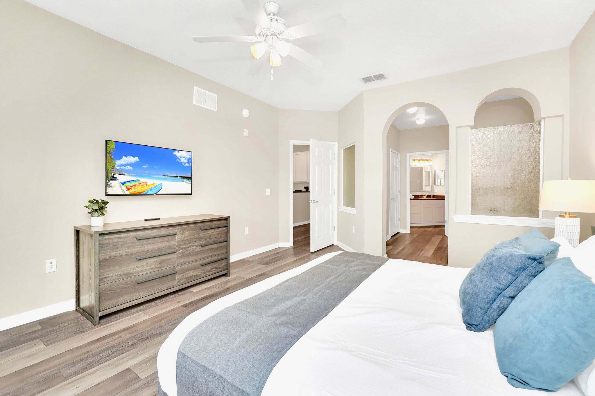 The spacious master bedroom features a large bed with crisp white linens, blue accent pillows, and a gray throw, complemented by light wood-look flooring. A wall-mounted TV sits above a rustic wooden dresser, and an arched entryway leads to the en-suite bathroom.