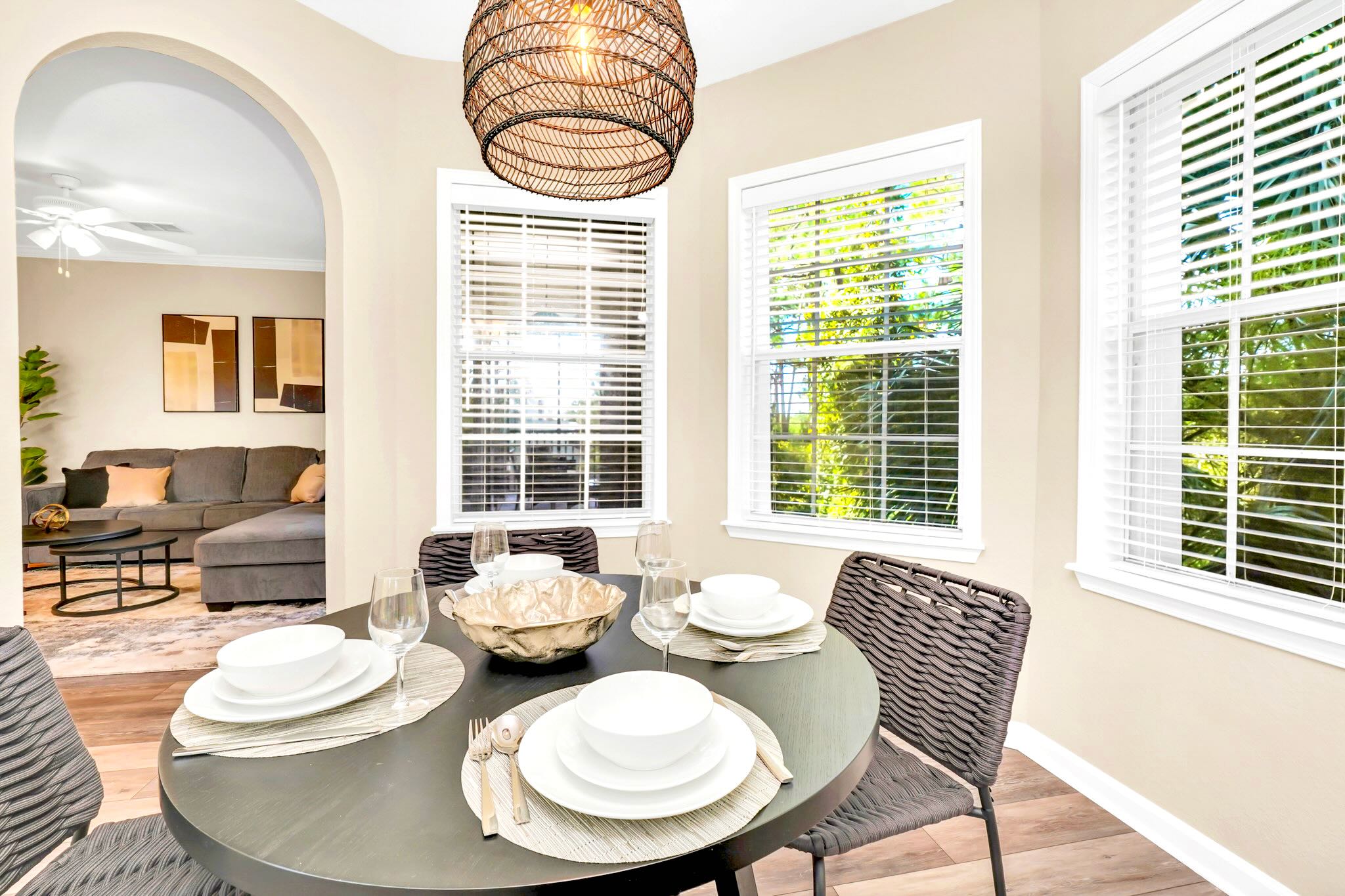 A cozy dining area features a round dark wood table set for four with white dishes and wine glasses, illuminated by a woven pendant light. Two windows with white blinds overlook lush greenery, and an arched doorway leads to the adjacent living space.