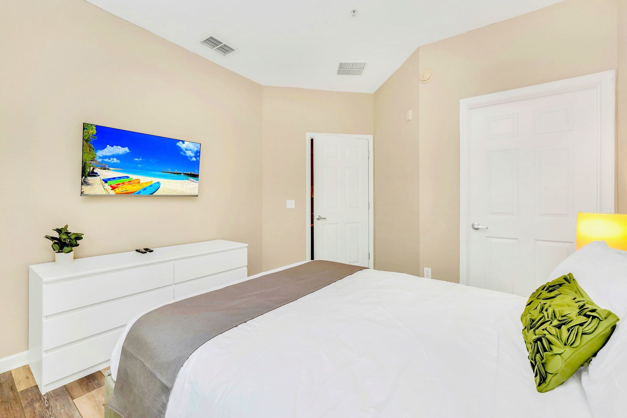 Another view of the second bedroom shows a wall-mounted TV positioned above a sleek white dresser, set against light beige walls and wood-look flooring. A closed white door is visible, and a green decorative pillow adds a pop of color to the bed.