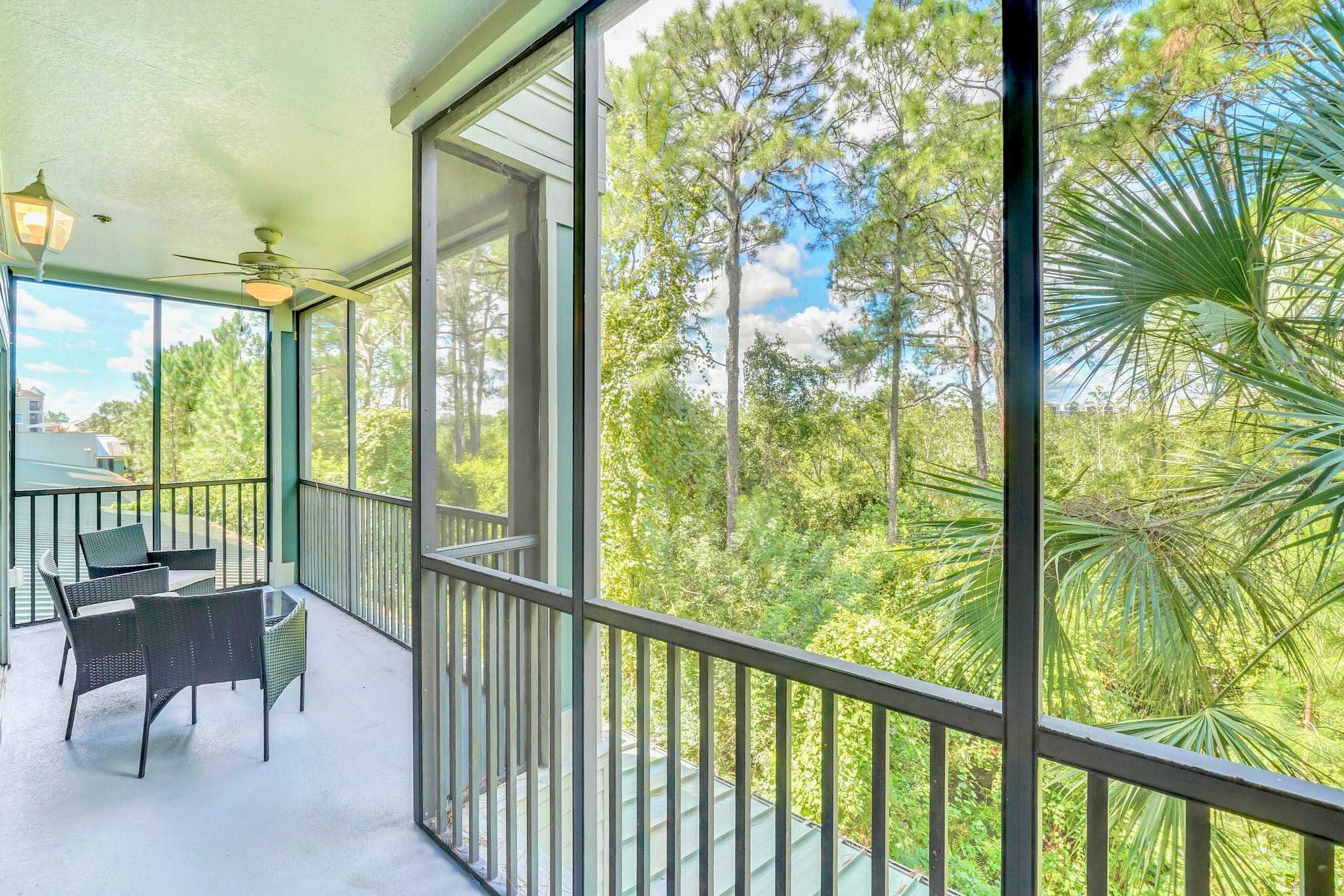 Screened balcony with dark wicker outdoor seating, including two chairs and a small table, offering a view of lush green trees and foliage, with a ceiling fan overhead.