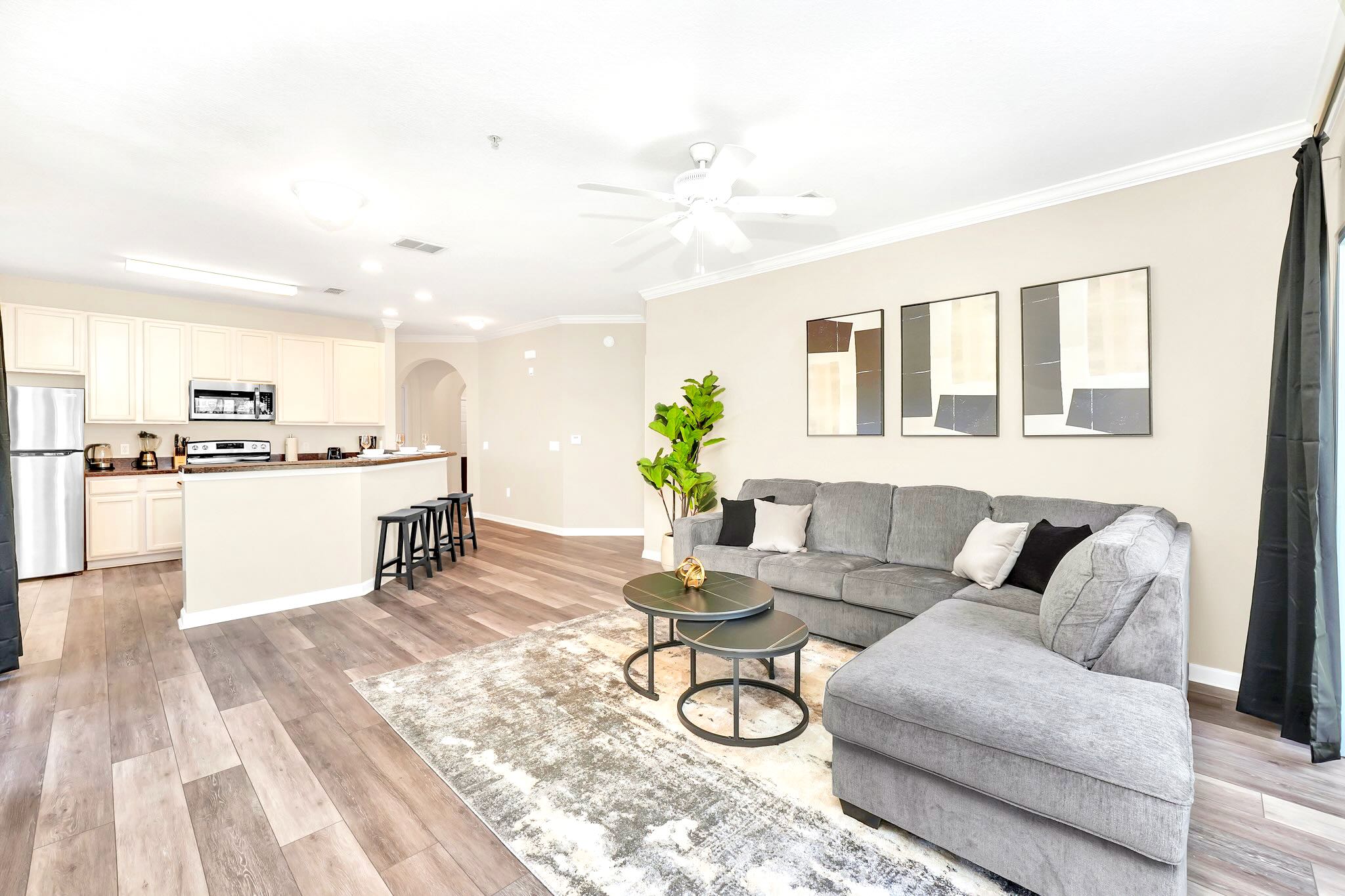 Open-concept living room with a gray sectional sofa, two round coffee tables on a patterned rug, and abstract wall art, flowing into a kitchen with a breakfast bar and light-colored cabinets.