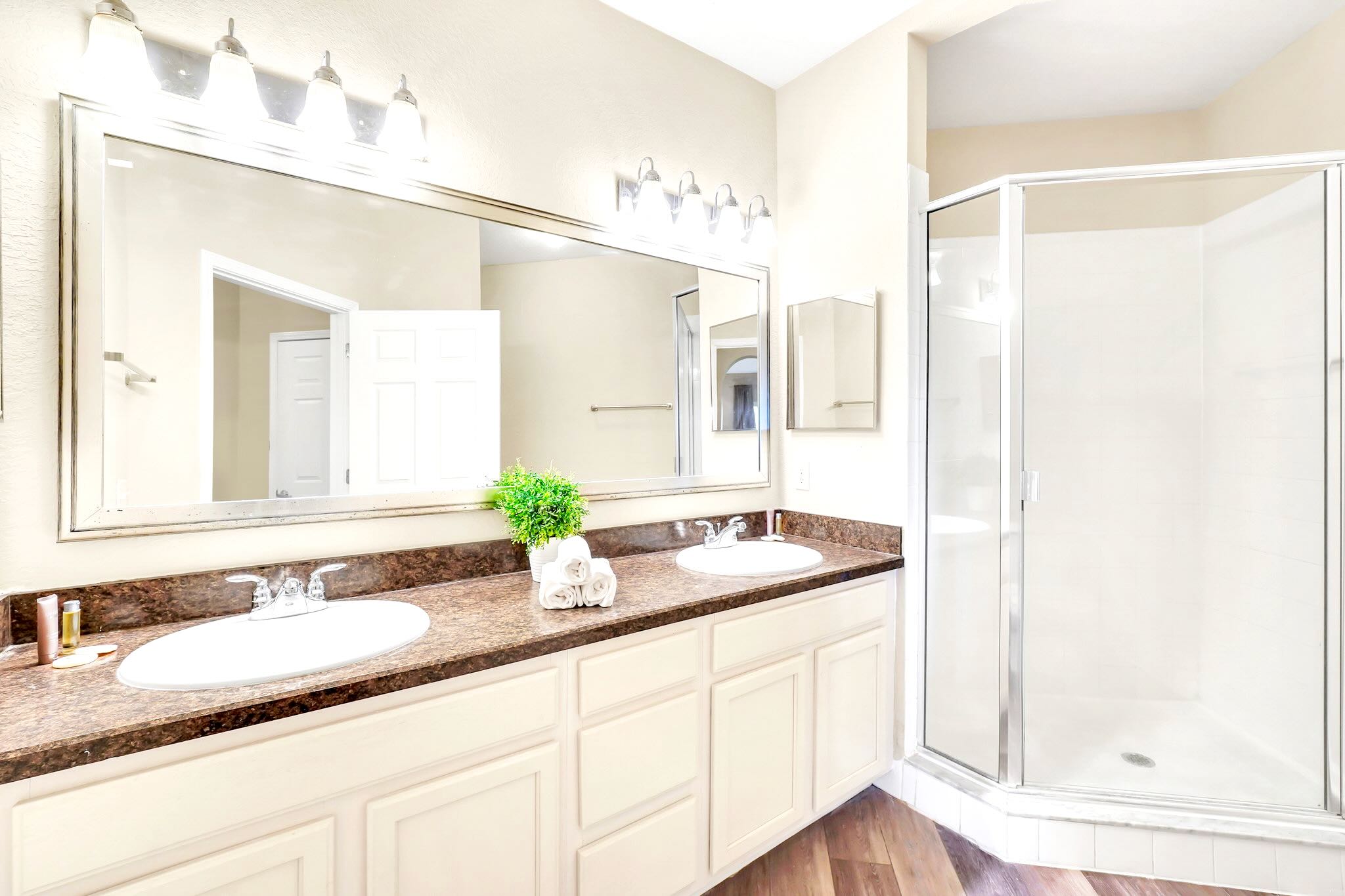 The master bathroom vanity boasts double sinks set in a dark speckled countertop, illuminated by a large mirror and bright overhead lighting. A glass-enclosed shower with a white base is visible to the right, completing the modern design.