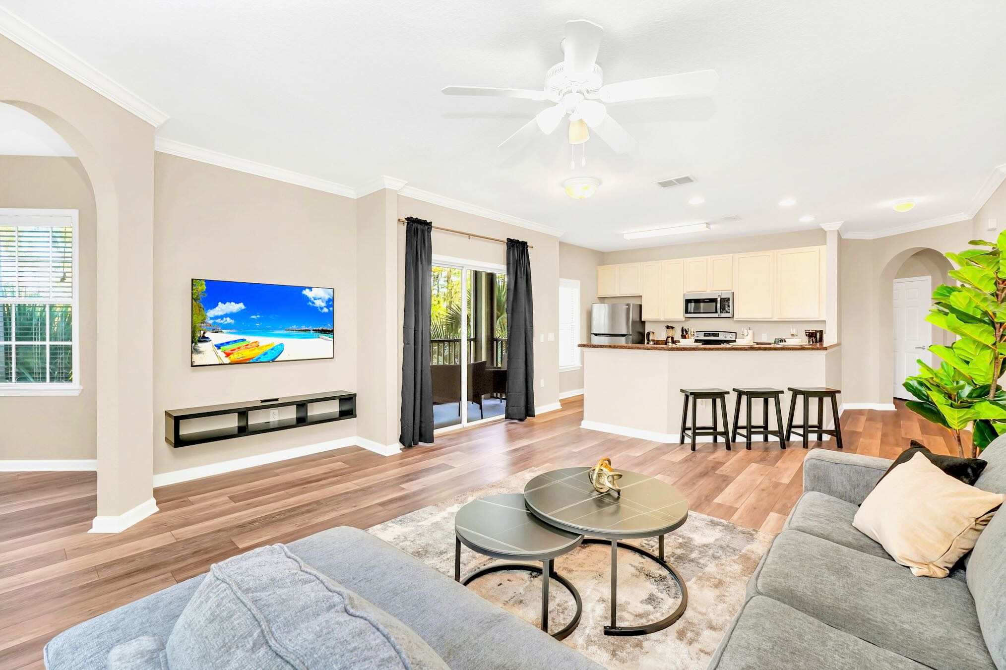 Modern living room with light wood-look flooring, a gray sectional sofa, two round coffee tables on a patterned rug, and a wall-mounted TV, open to a kitchen with a breakfast bar and light cabinetry.