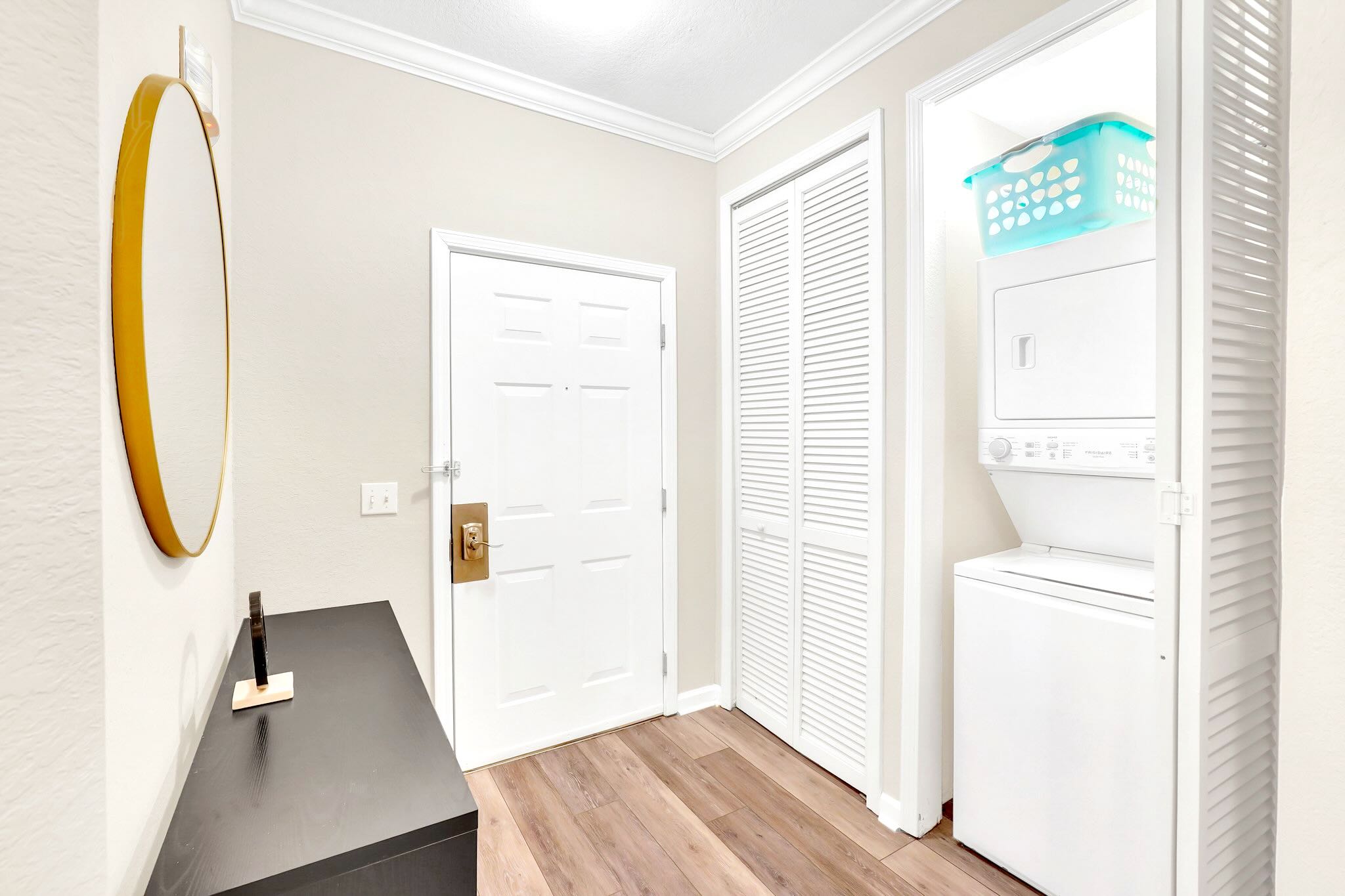 A bright foyer features light wood-look flooring, a white front door with a brass handle, and a stacked washer and dryer unit neatly tucked into a louvered closet. A round gold-framed mirror hangs above a dark console table.