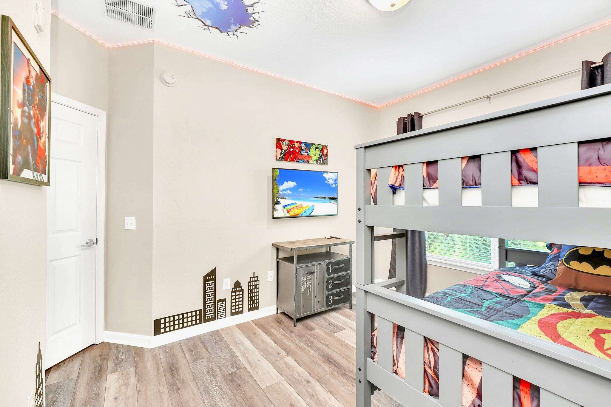 A vibrant kids' bedroom features a grey bunk bed with superhero bedding, light wood-look flooring, and light beige walls adorned with city skyline decals and a wall-mounted TV. Red LED strip lights line the ceiling, adding a playful touch.