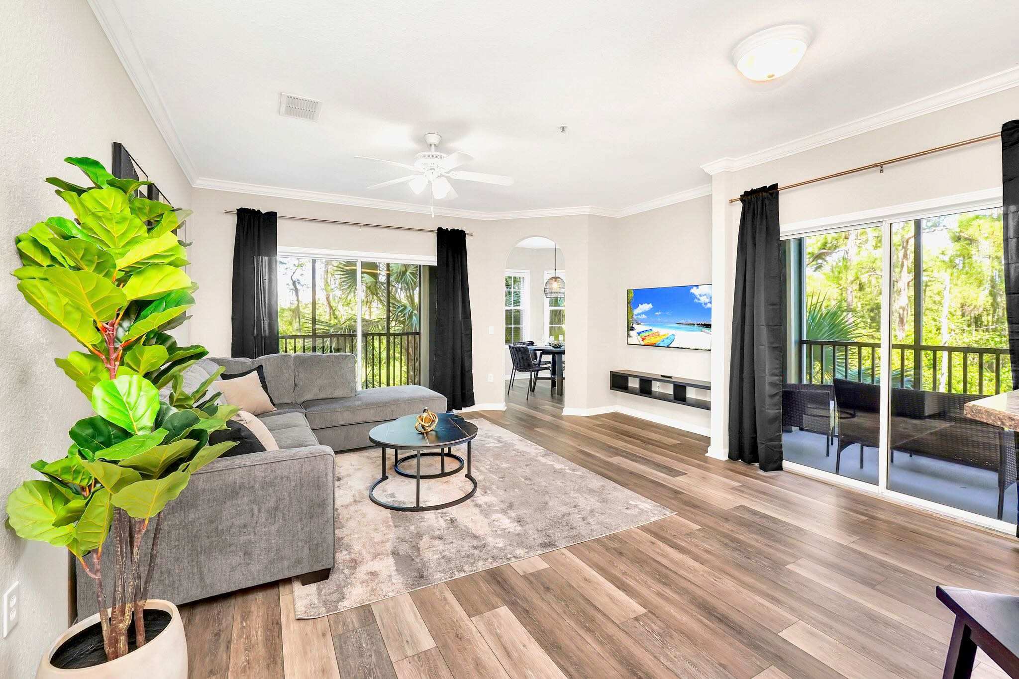 Wide view of a modern living room with a gray sectional sofa, two round coffee tables on a patterned rug, a large potted plant, and two glass sliding doors leading to separate balconies.