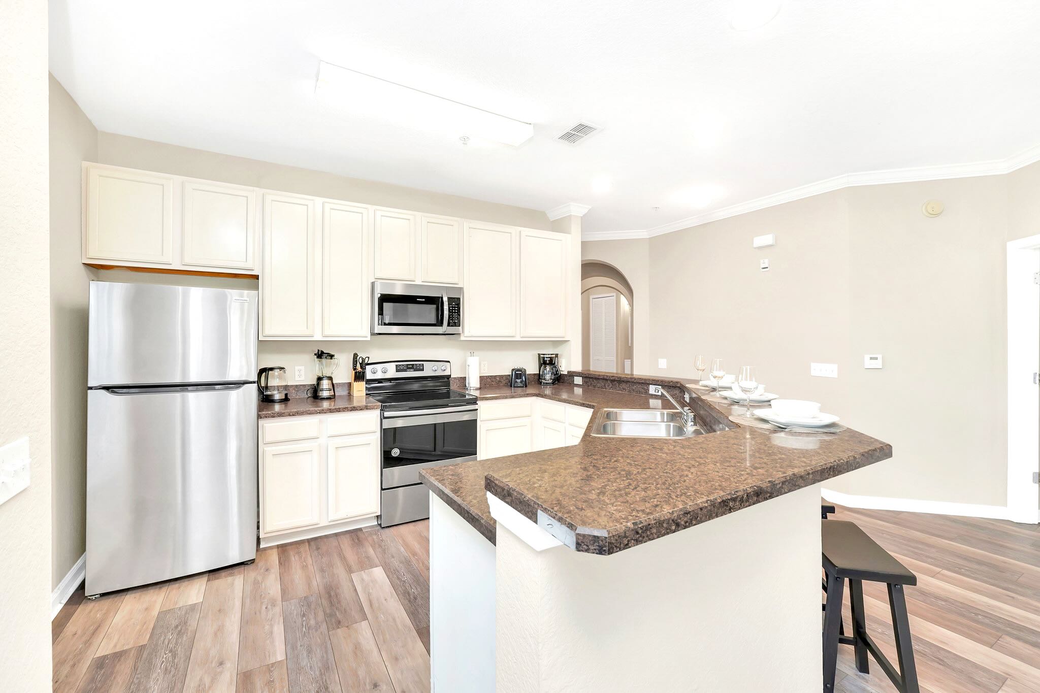 A well-equipped kitchen showcases light cream shaker-style cabinets, dark speckled countertops, and a suite of stainless steel appliances including a refrigerator, microwave, and electric range. A double sink is integrated into the breakfast bar, complementing the warm wood-look flooring.