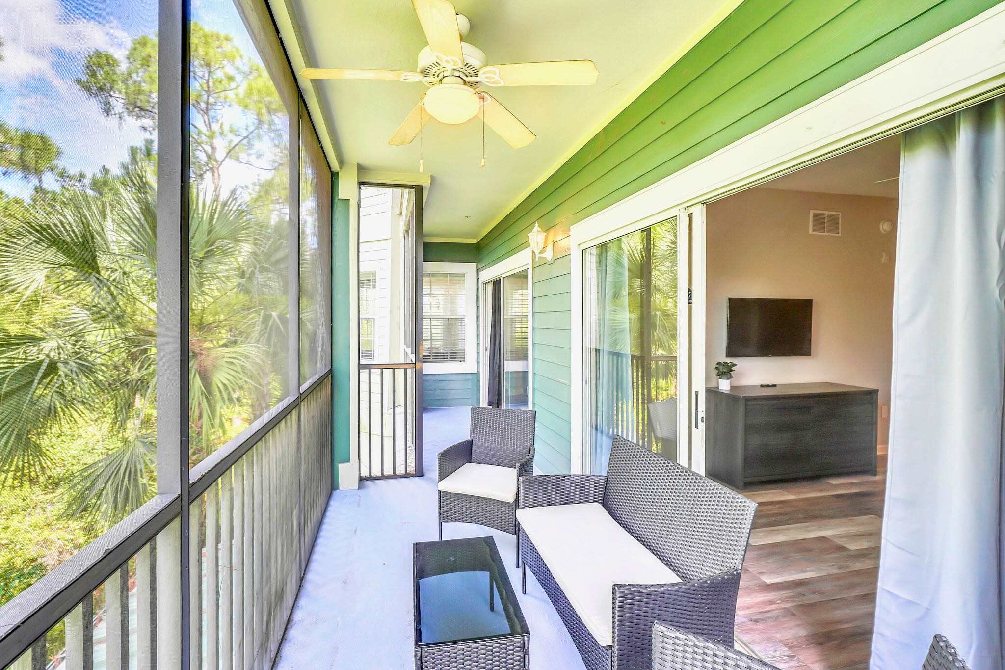Long screened balcony with a vibrant green accent wall, furnished with dark wicker outdoor seating including a sofa and chairs, overlooking lush trees and foliage.