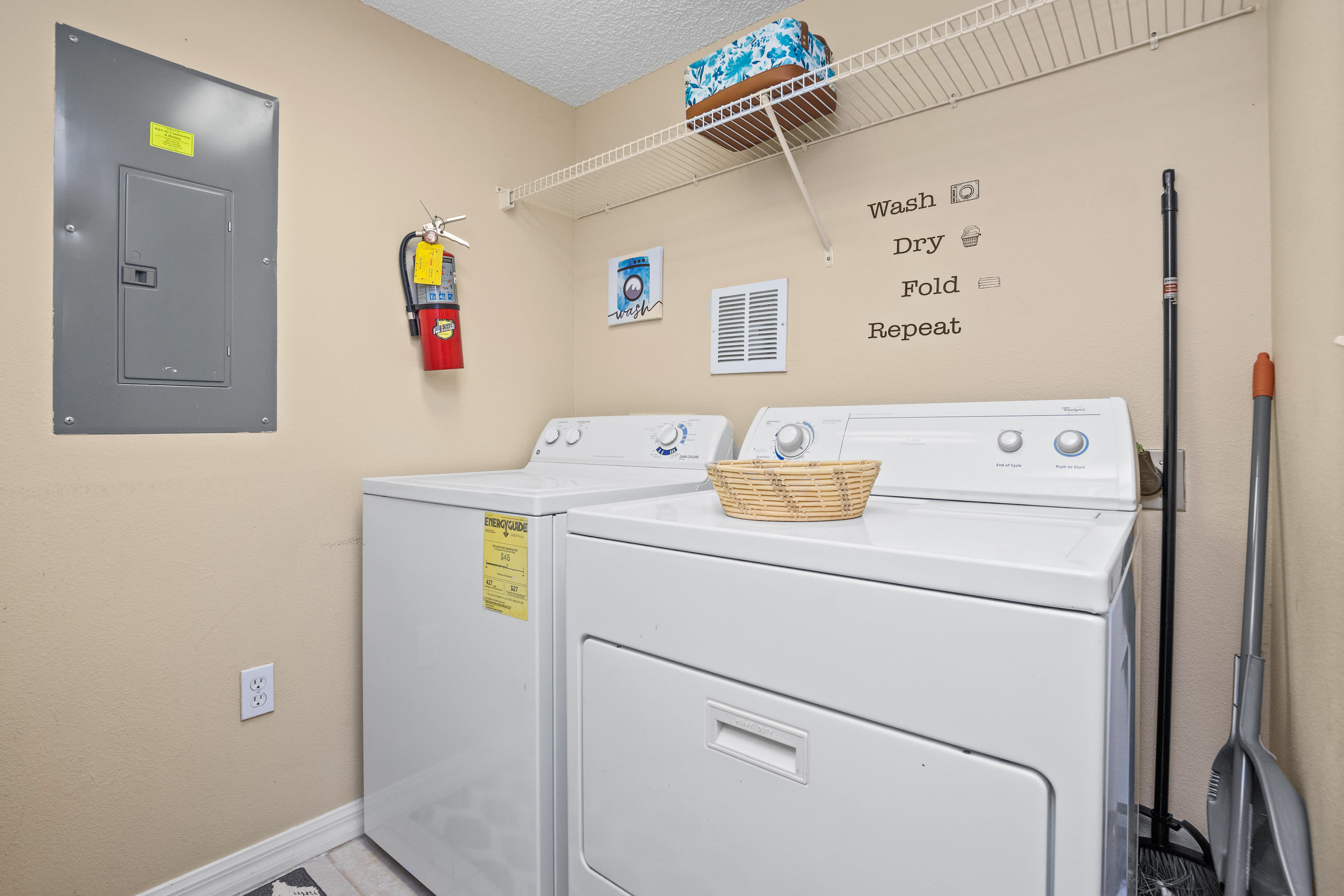Functional laundry room equipped with a white top-loading washing machine and dryer, a wire shelf above, and a fire extinguisher mounted on the wall. A 'Wash Dry Fold Repeat' decal adds a touch of decor to the light yellow wall.