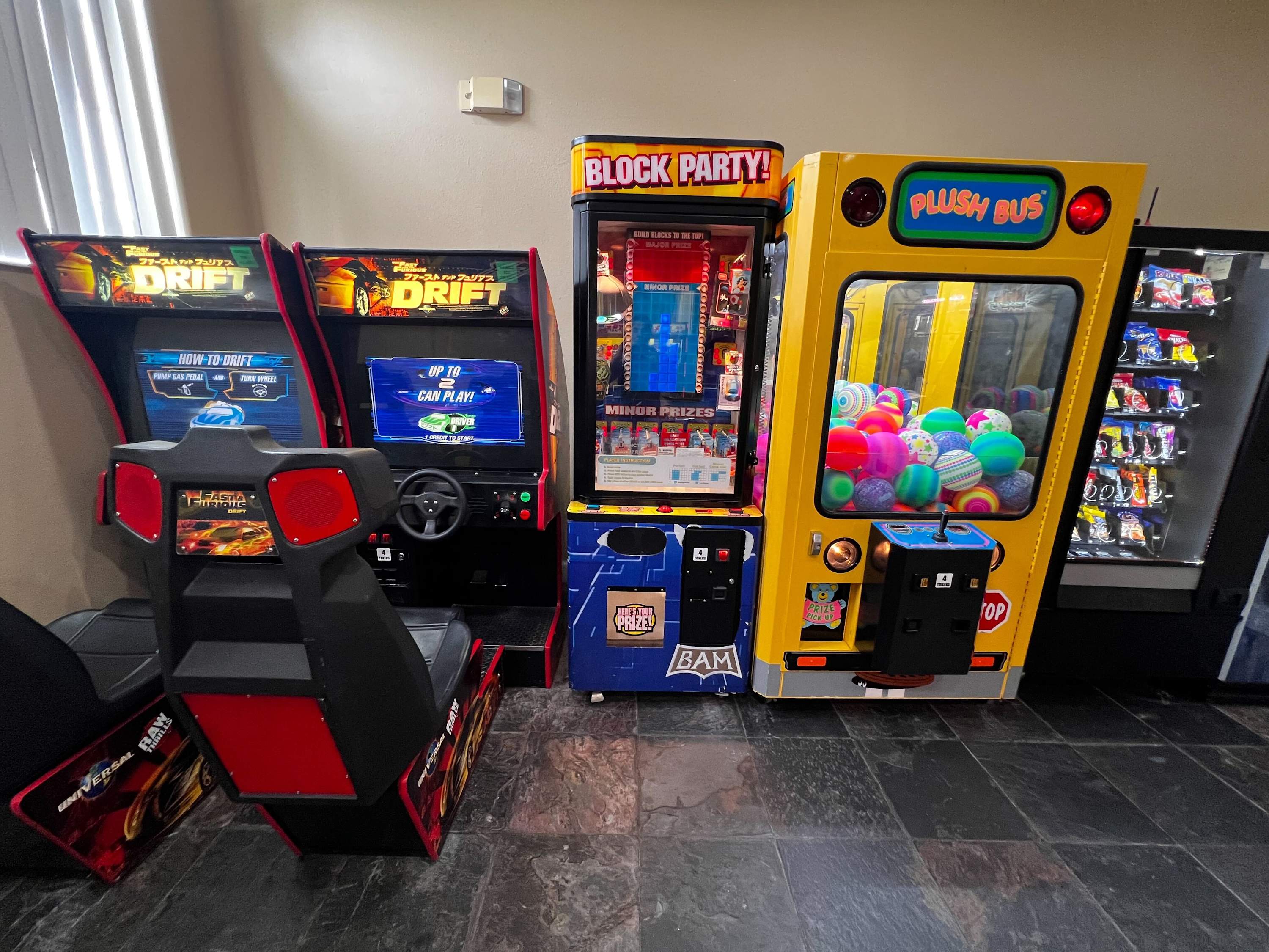A section of a game room features several arcade machines, including two racing simulators, a "Block Party" prize game, and a "Plush Bus" claw machine filled with colorful plush toys. A snack vending machine is also present.