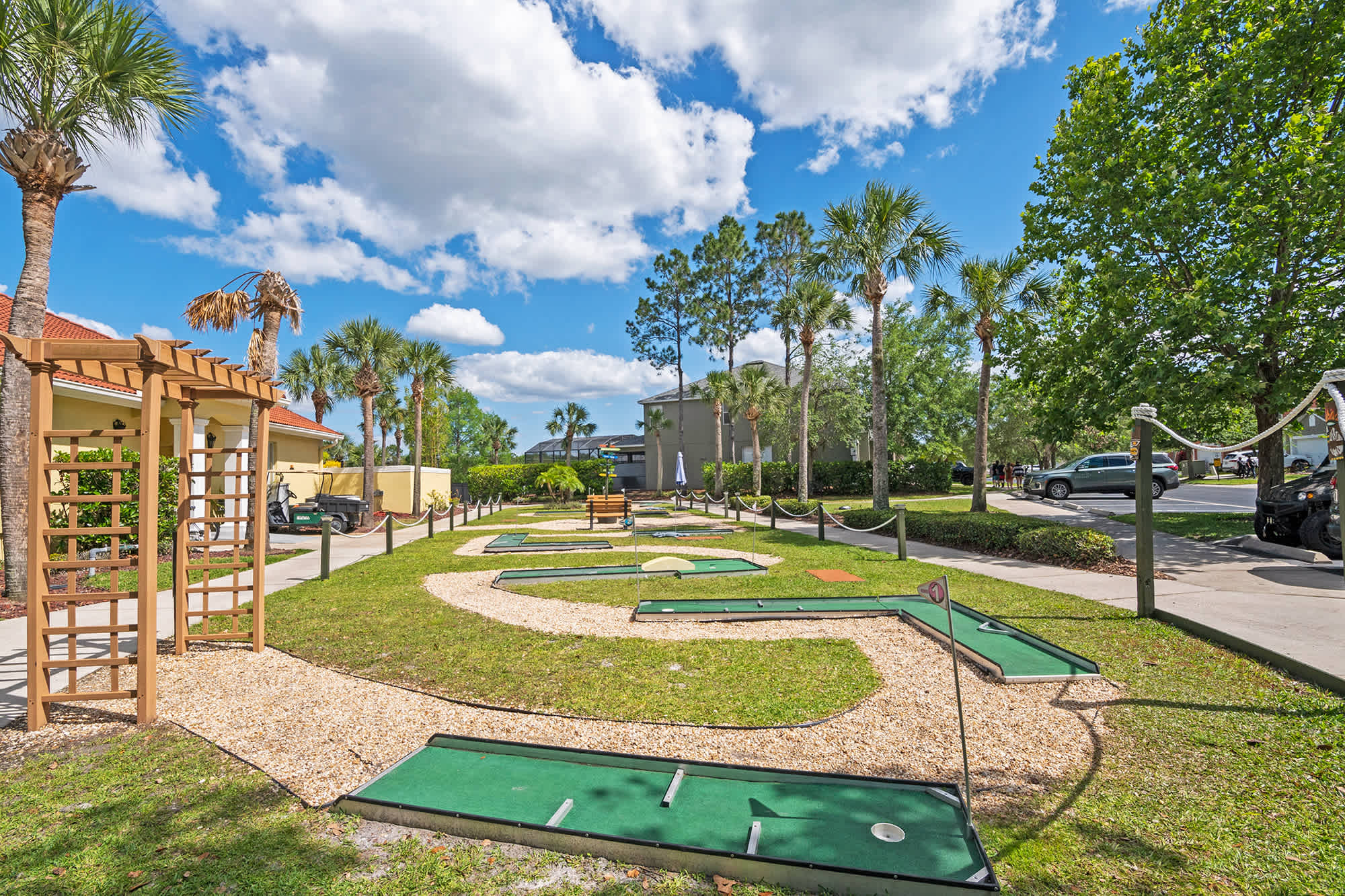 A vibrant outdoor mini-golf course features multiple green putting areas connected by light-colored gravel paths, surrounded by lush green grass and tall palm trees under a bright blue sky. A wooden pergola structure is visible on the left.