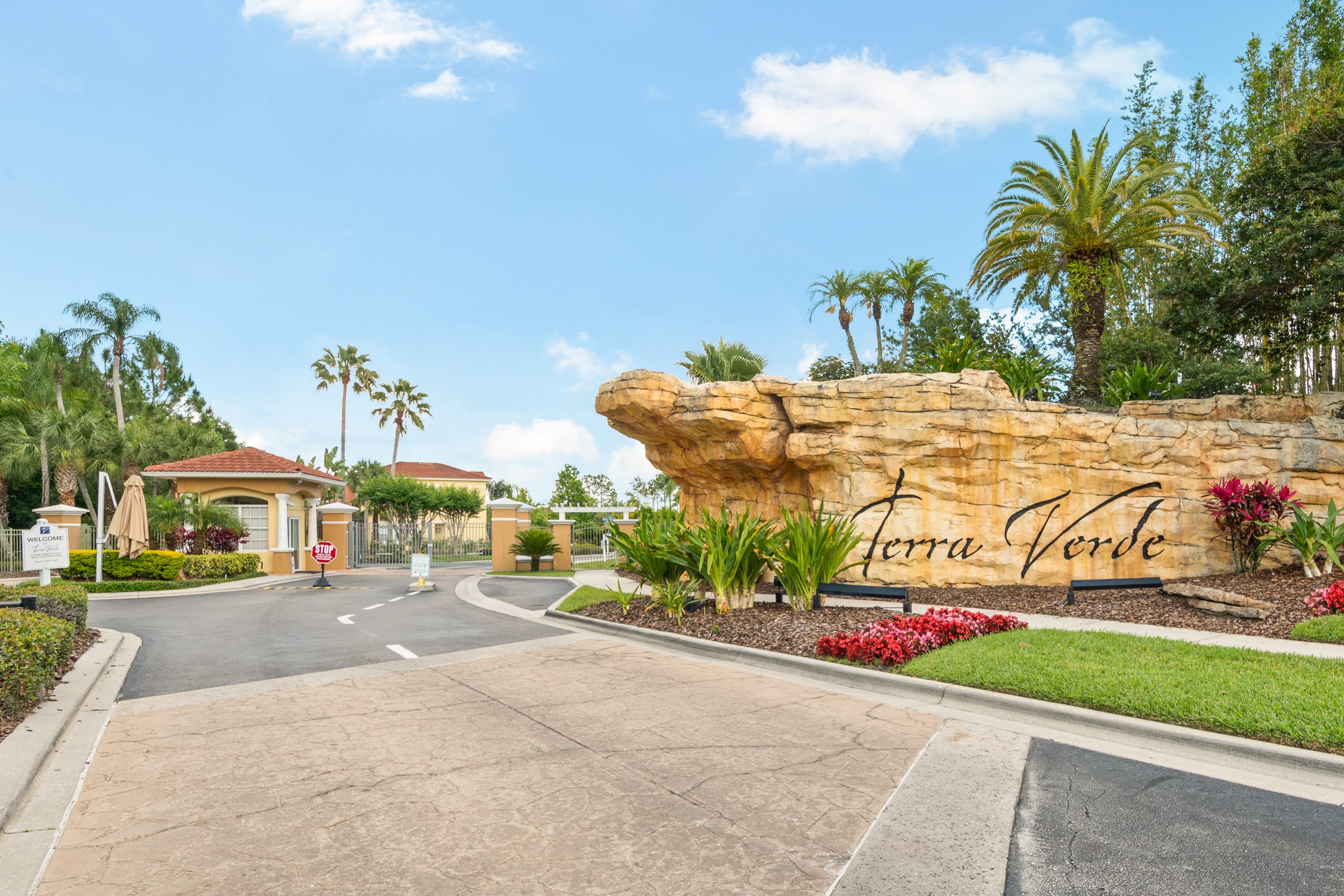 Impressive gated entrance to the 'Terra Verde' community, marked by a large, natural rock sign with the community name. The entrance features lush tropical landscaping, tall palm trees, and a security booth under a bright blue sky.