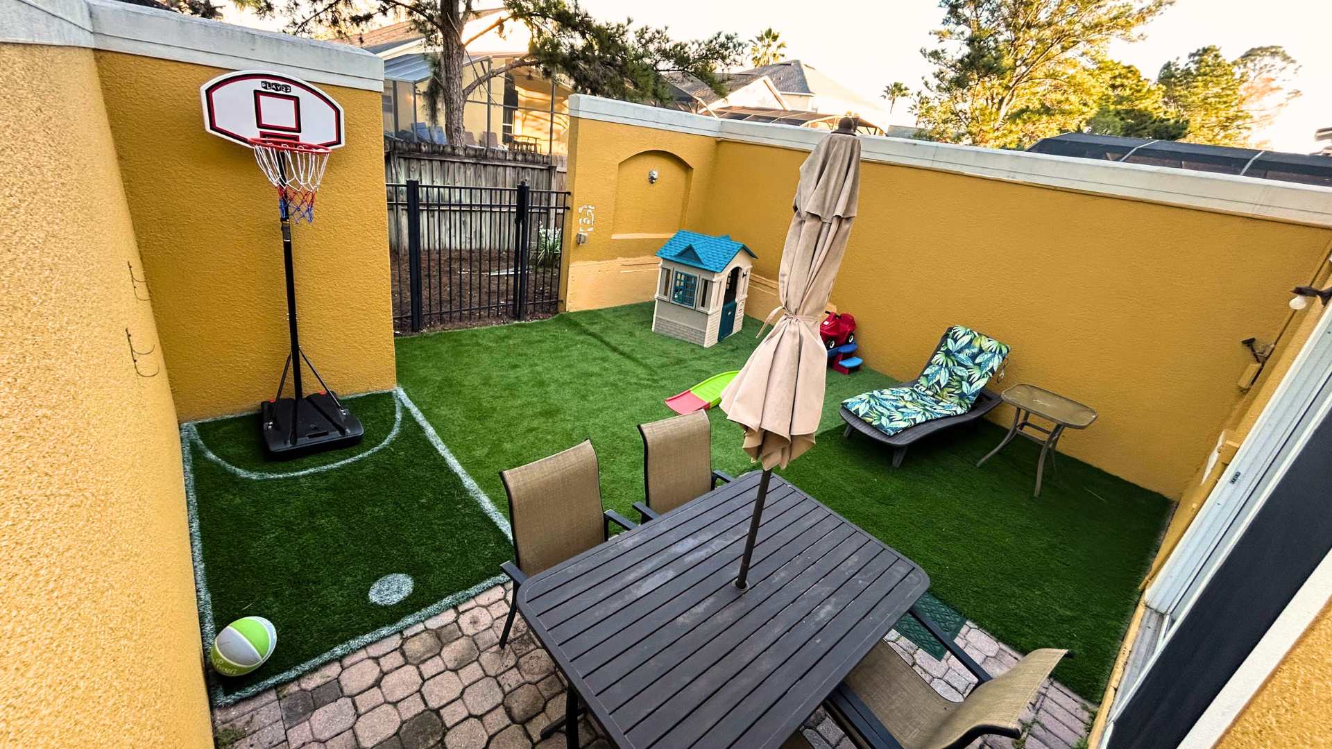 Enclosed outdoor patio with artificial turf, a basketball hoop, and a paved area with an outdoor dining table and umbrella. A small playhouse and lounge chair are also visible, creating a family-friendly outdoor space.