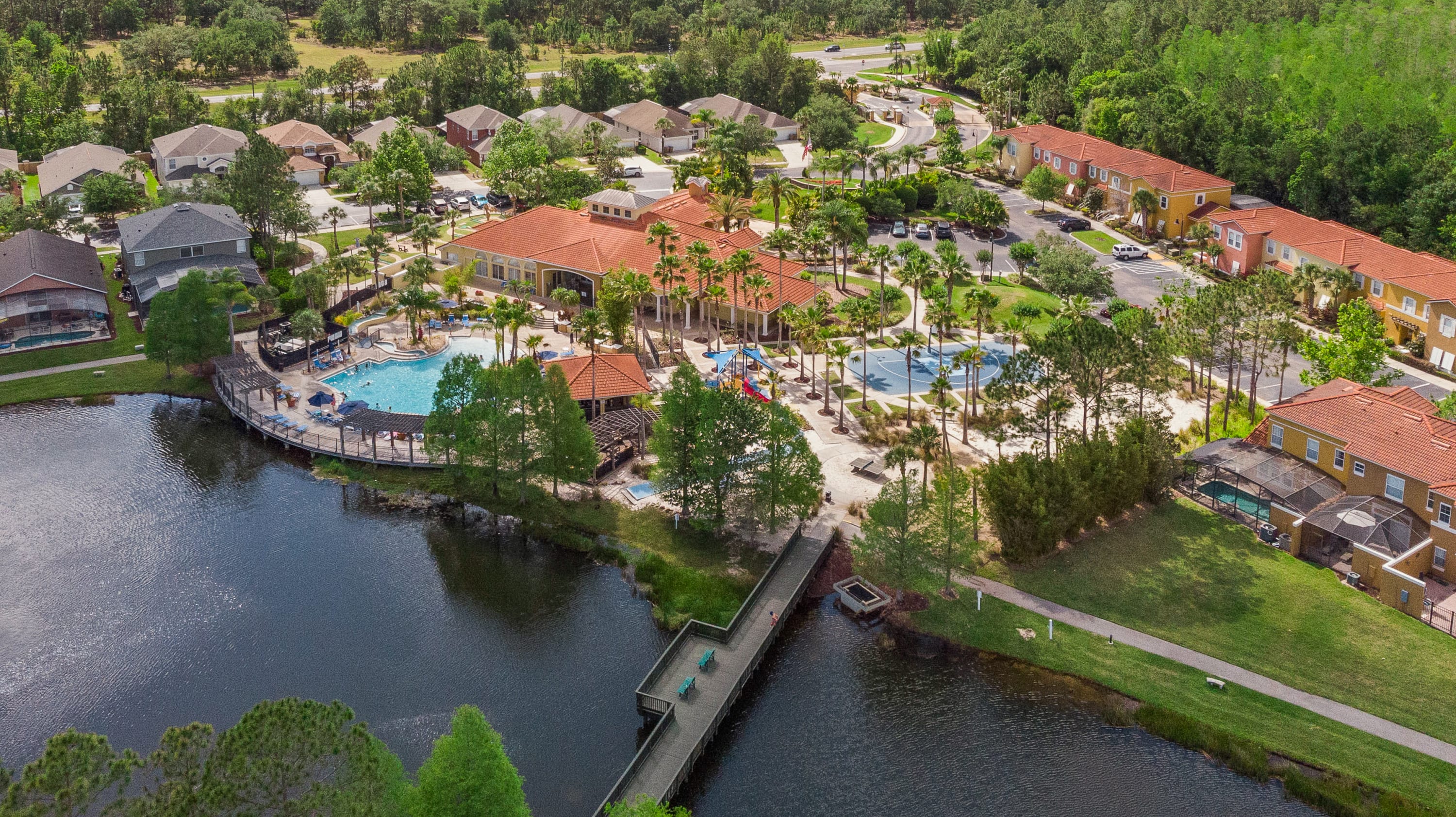 Aerial view of a resort community featuring a large communal pool area with lounge chairs, a basketball court, and a clubhouse. The property is surrounded by lush greenery, a lake, and residential buildings with red tile roofs.