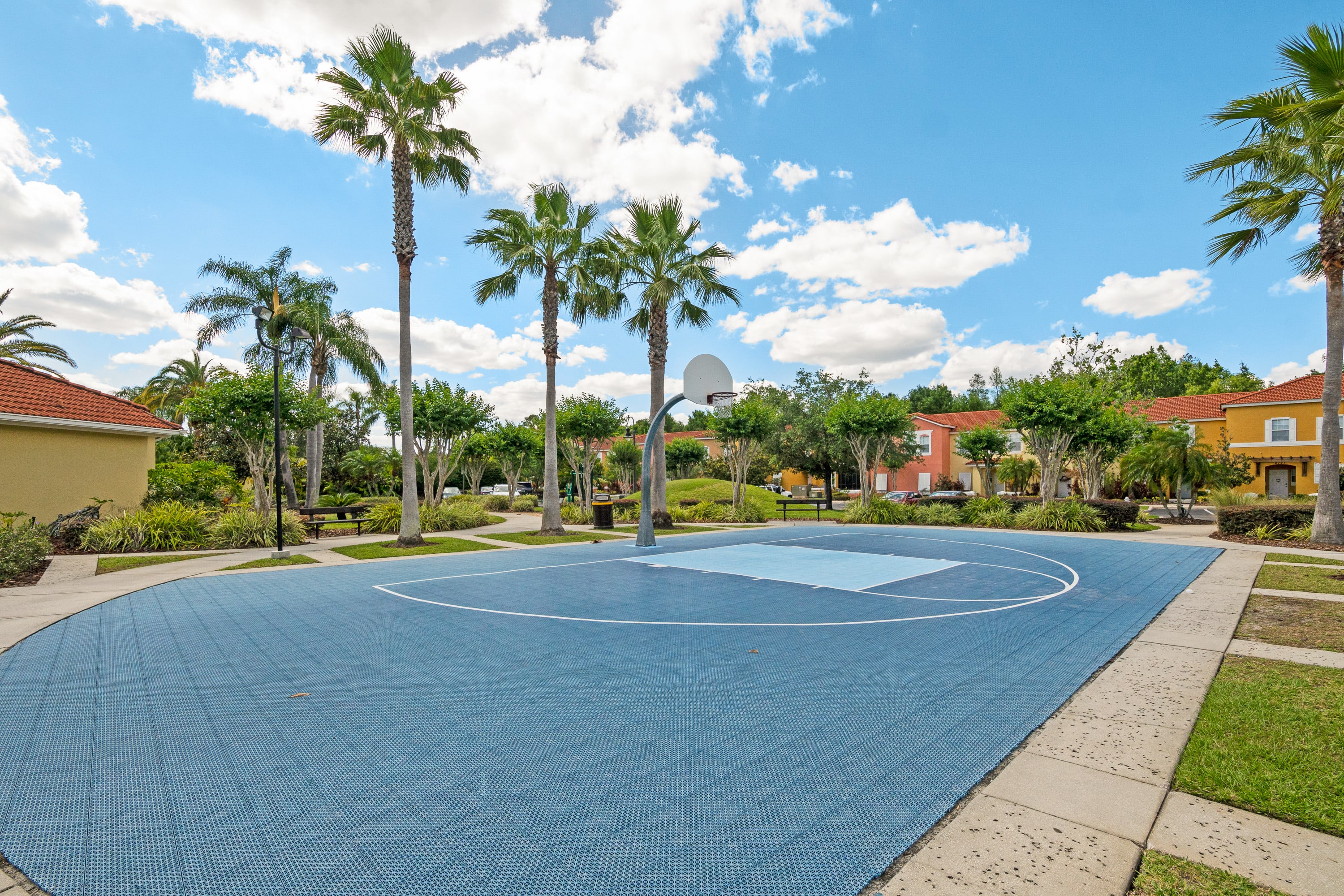 A bright blue outdoor basketball court with white markings and a single hoop is set amidst green grass and tall palm trees. Resort buildings with orange tiled roofs are visible in the background under a clear blue sky.