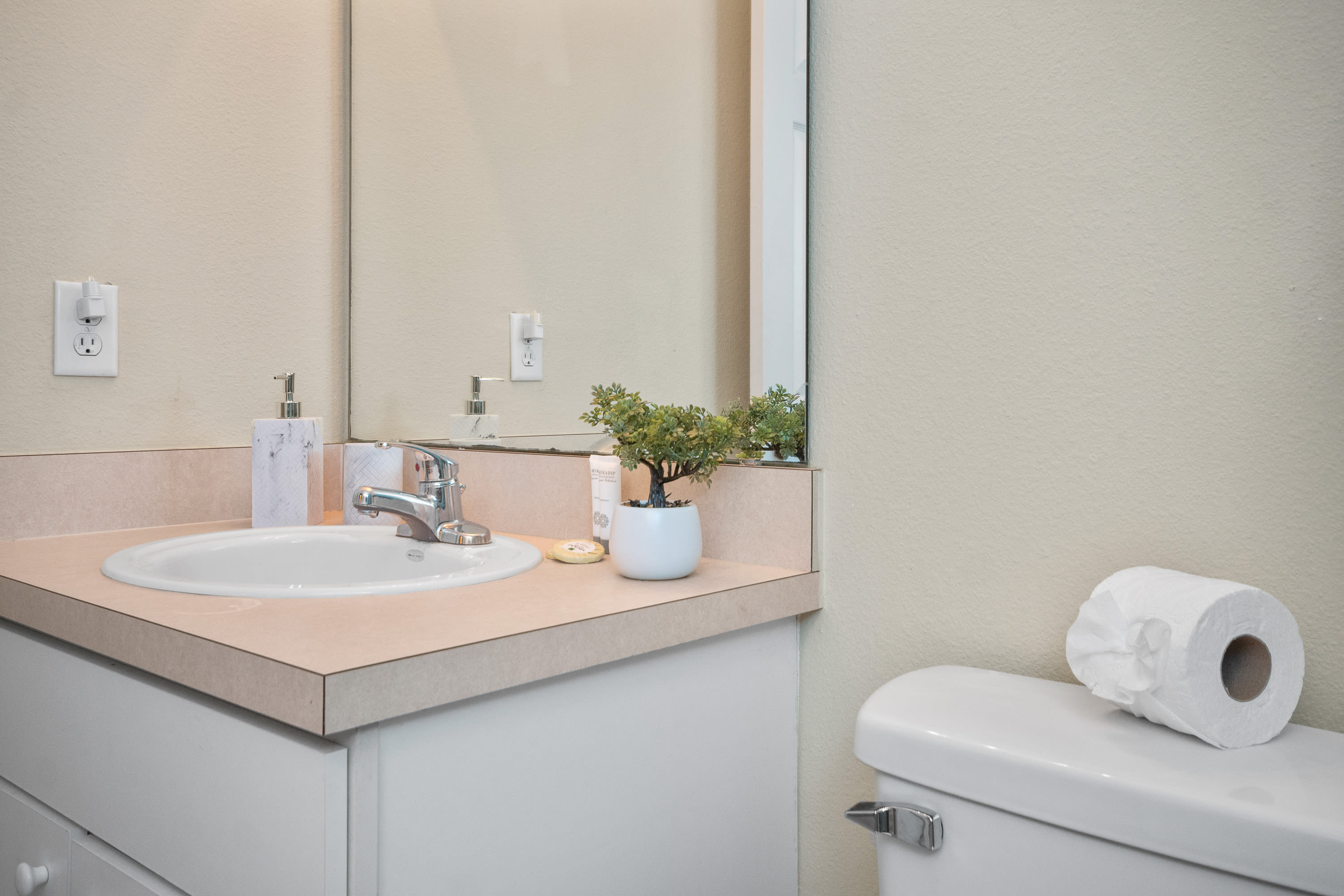 Close-up of a bathroom vanity with a white oval sink, chrome faucet, peach countertop, and white shaker-style cabinets. A small potted plant and marble-patterned soap dispenser adorn the counter, with a toilet nearby.