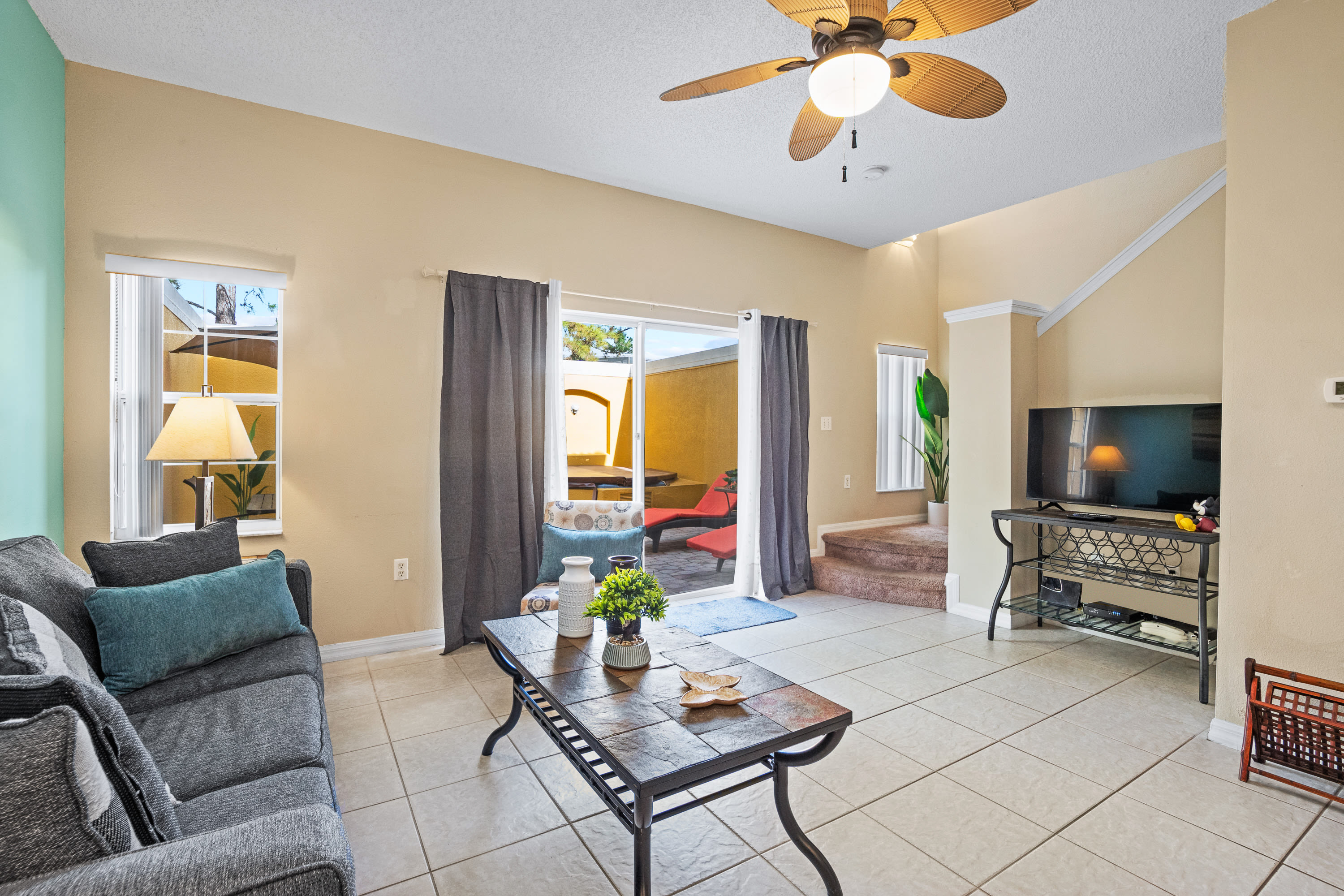 Living room with a gray sofa, a slate-top coffee table, and a TV on a black console. Sliding glass doors open to an outdoor patio, and a ceiling fan provides air circulation in the tiled space.