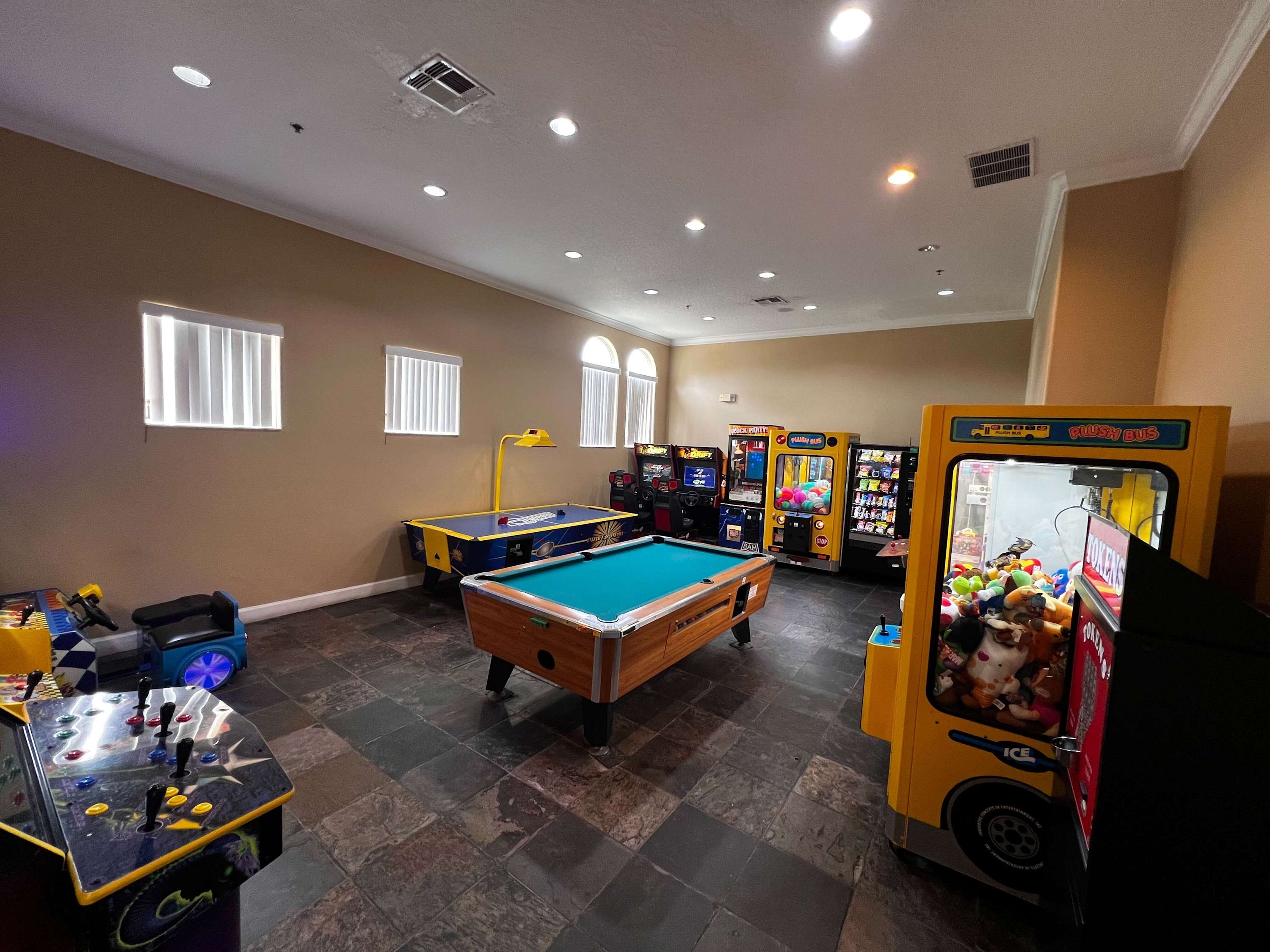 A spacious game room features a central pool table, an air hockey table, and multiple arcade machines, including claw games, set on a dark tiled floor. Light-colored walls and arched windows provide natural light.