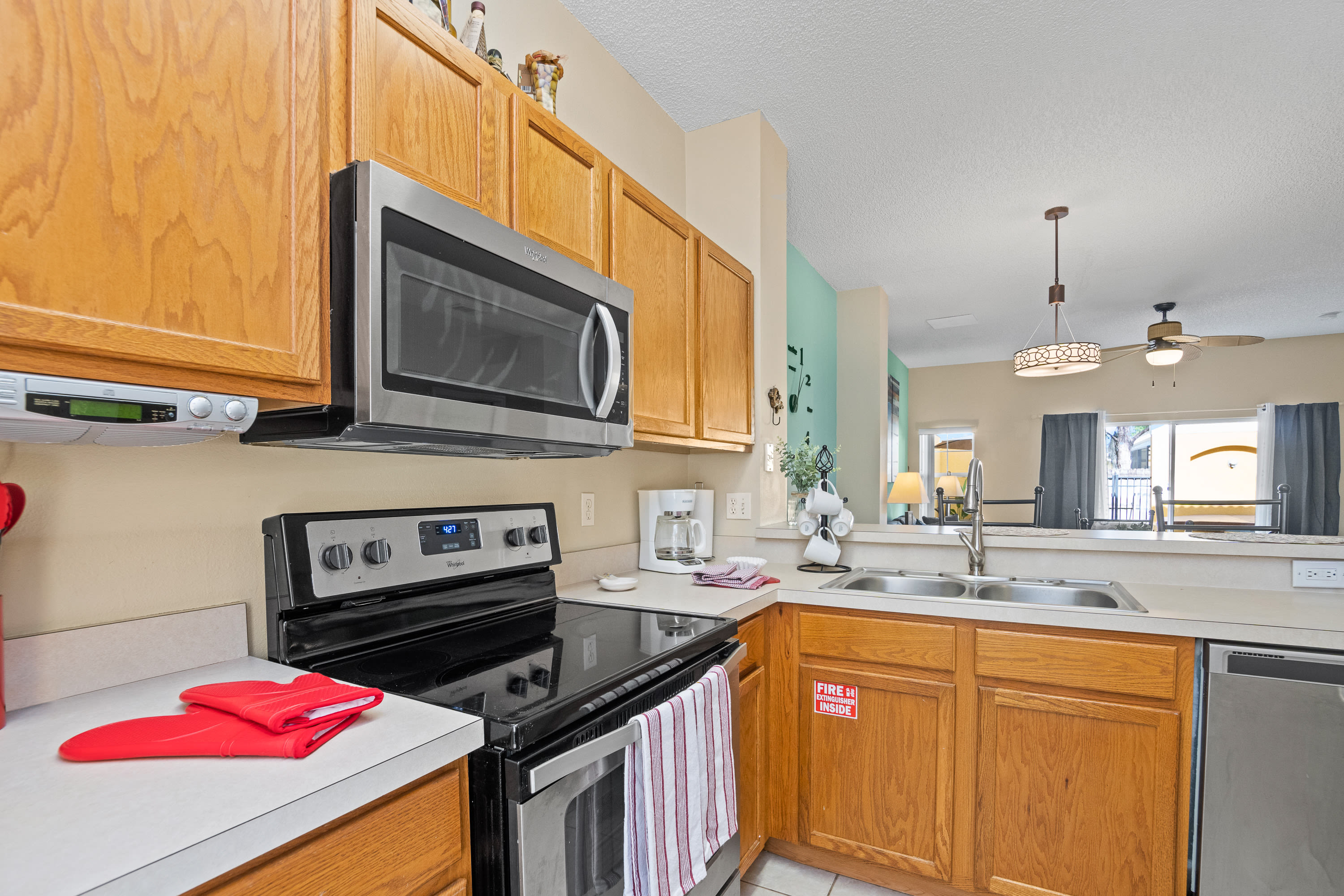 A functional kitchen featuring a black electric range with an overhead stainless steel microwave, light wood cabinetry, and white countertops.