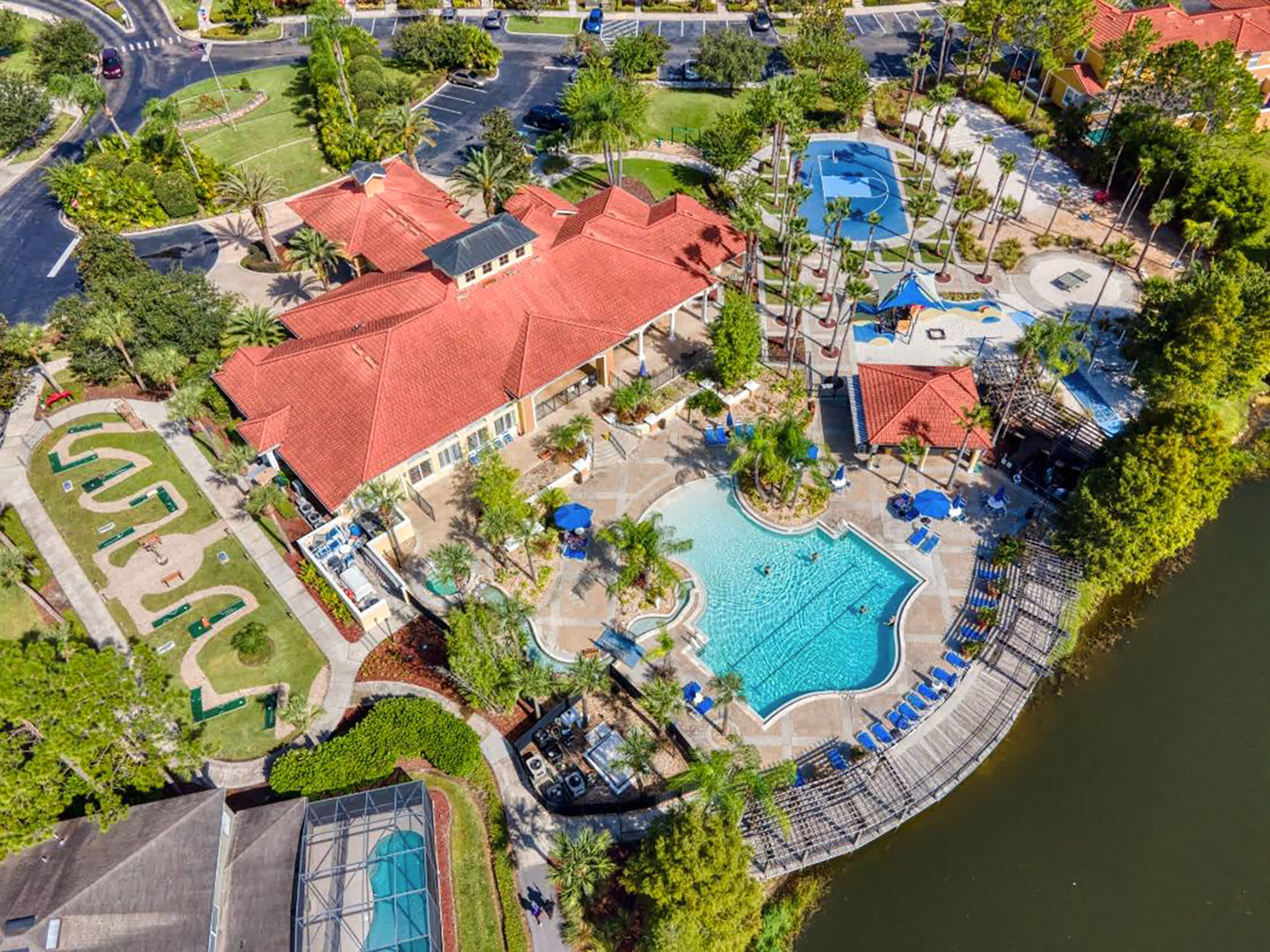 Expansive aerial view of a vibrant resort pool complex, featuring a large freeform swimming pool, a separate splash pad, a miniature golf course, and a clubhouse with a red tile roof, all surrounded by abundant palm trees and greenery.