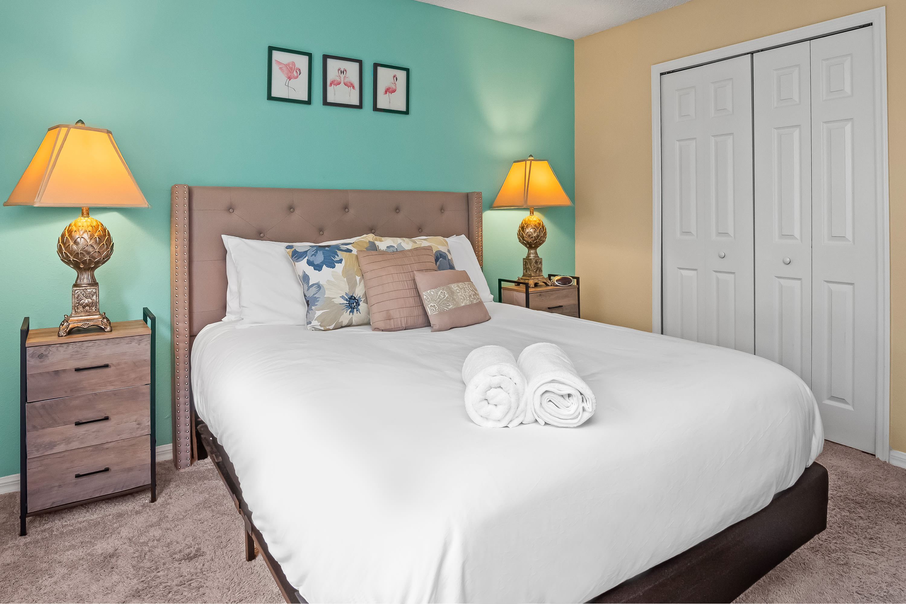 A cozy bedroom with a queen-sized bed featuring a tufted headboard, two wooden nightstands with lamps, and a vibrant teal accent wall.