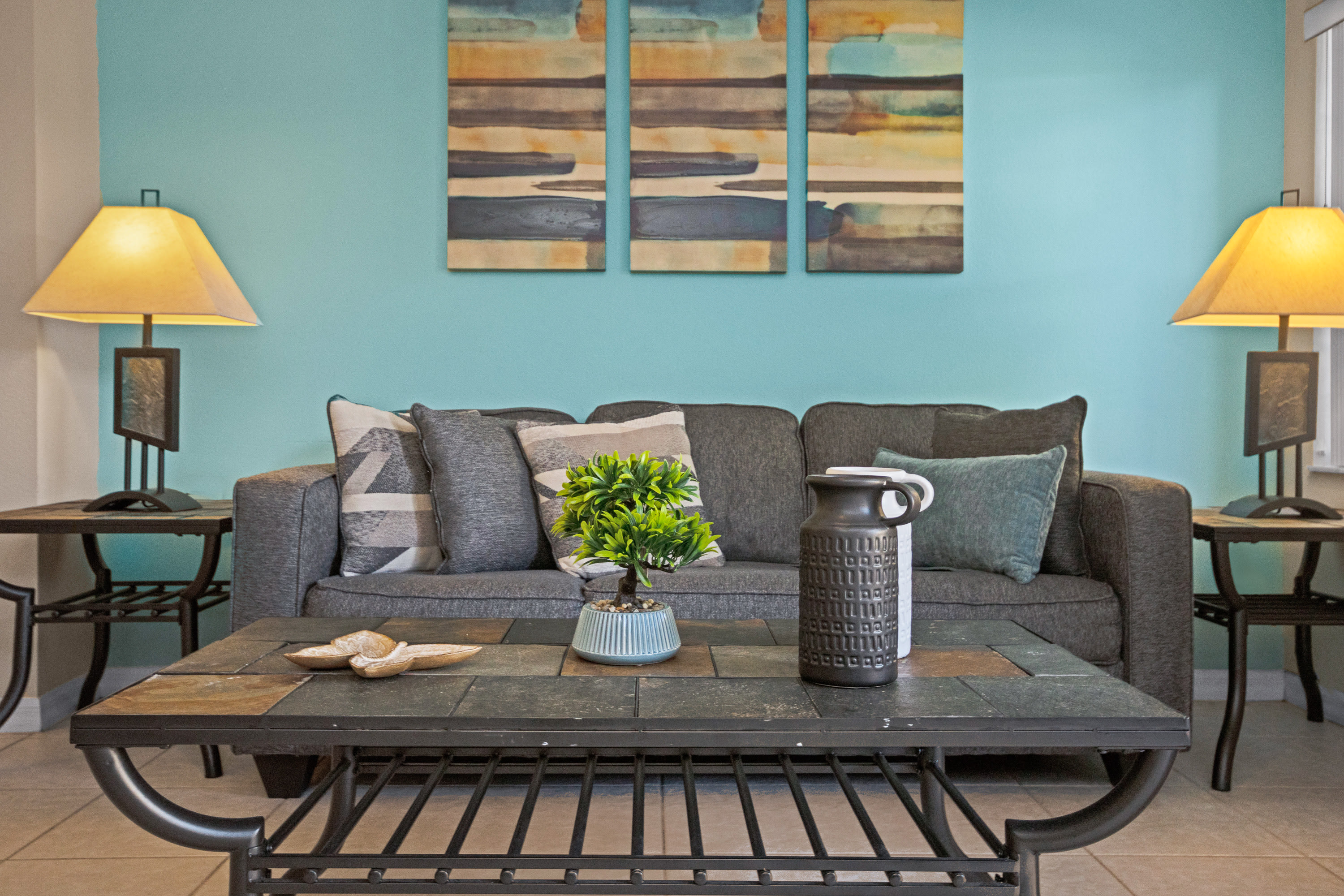 Close-up of a gray sofa adorned with throw pillows, a slate-top coffee table with decorative items, and two end tables with lamps. A teal accent wall with abstract art is visible in the background.
