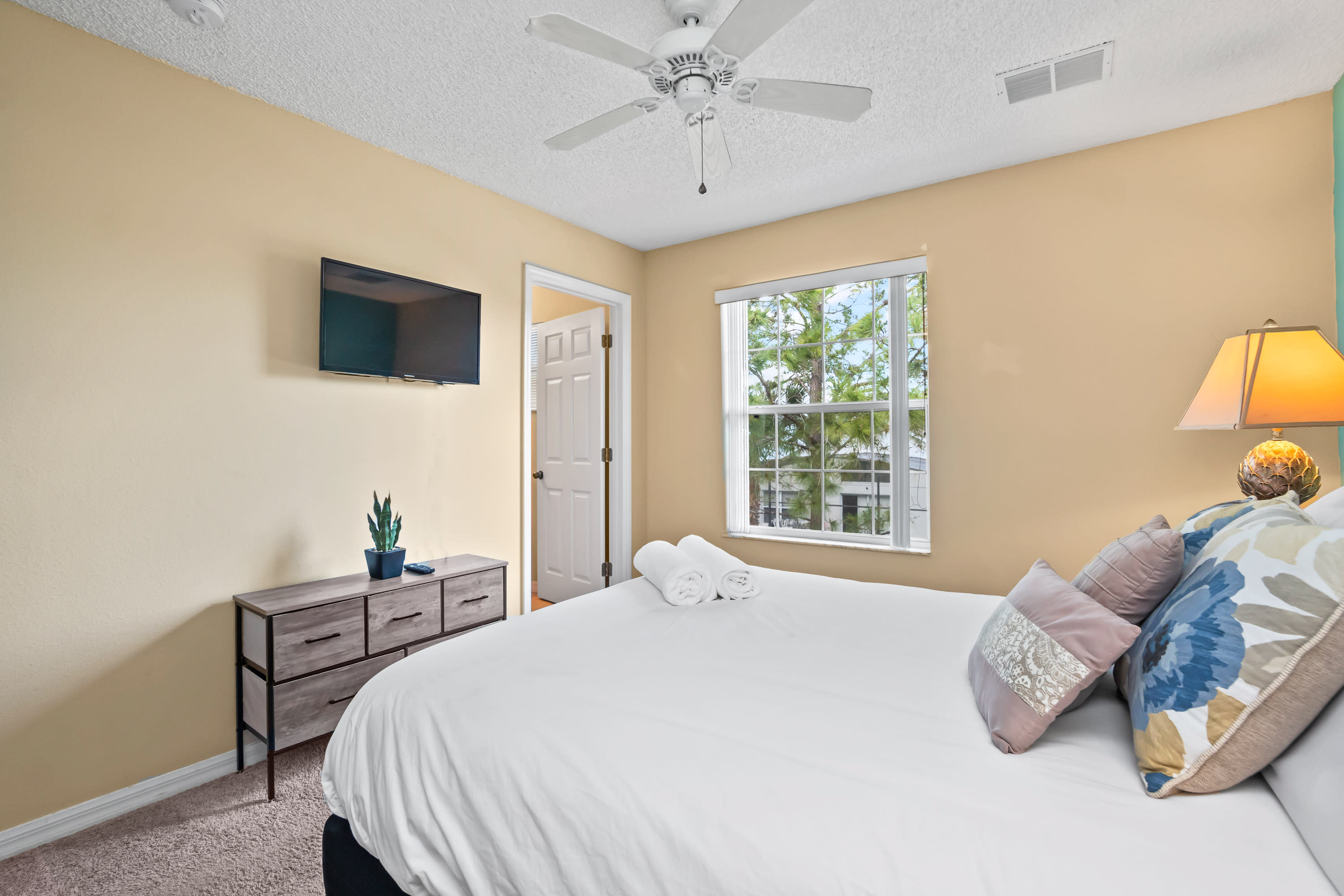 Another view of the bedroom showing the queen bed with decorative pillows, a wall-mounted TV above a dresser, and a window with blinds.