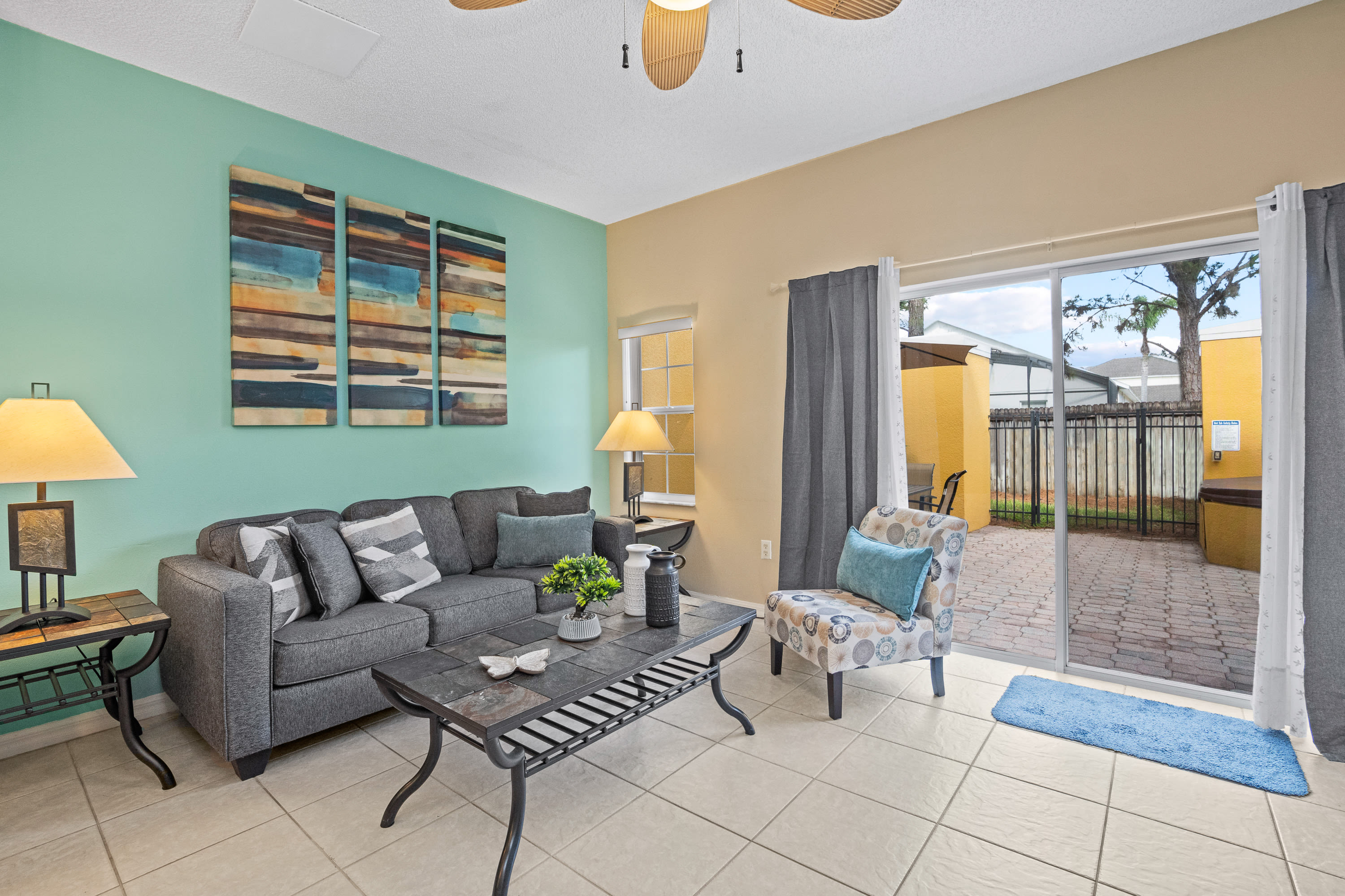 Bright living room with a gray sofa, a patterned accent chair, and a slate-top coffee table. Sliding glass doors lead to an outdoor patio, and a teal accent wall adds a pop of color.
