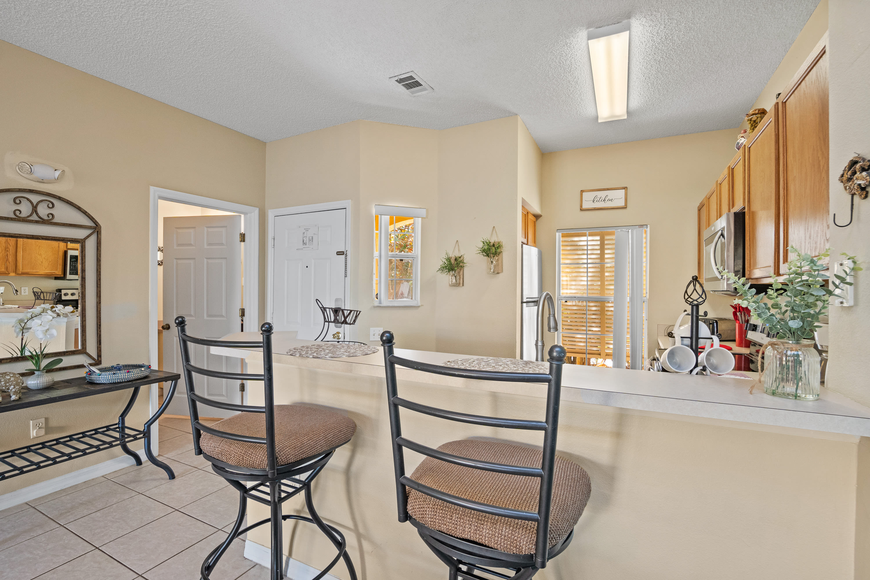 A bright kitchen breakfast bar with two wrought-iron bar stools, a white countertop, and a view into the kitchen with light wood cabinets and appliances.