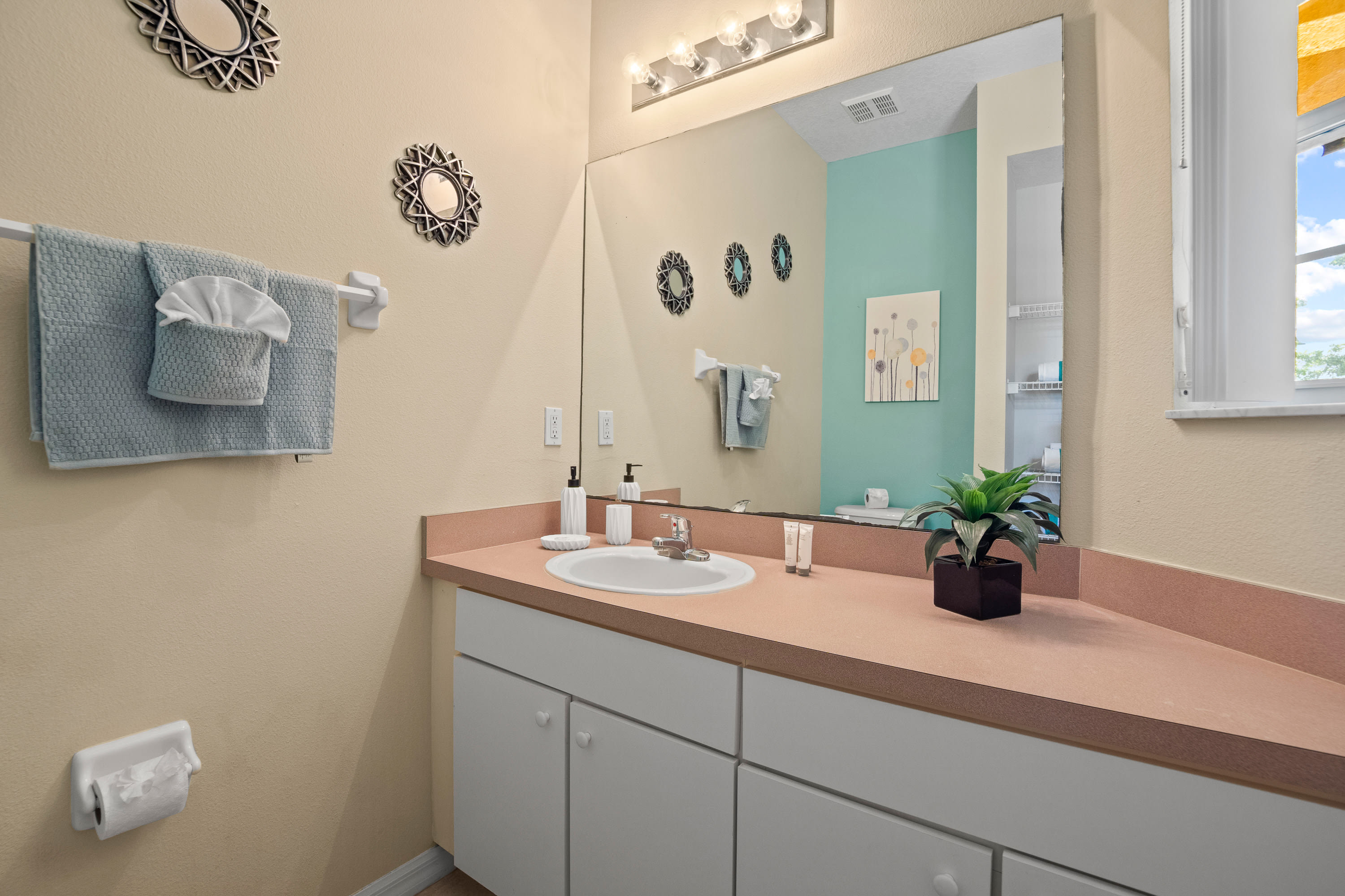 Bright bathroom featuring a single white sink set into a peach-colored countertop, white vanity cabinets, a large mirror, and a three-bulb light fixture. Light yellow walls are adorned with small, ornate decorative mirrors.