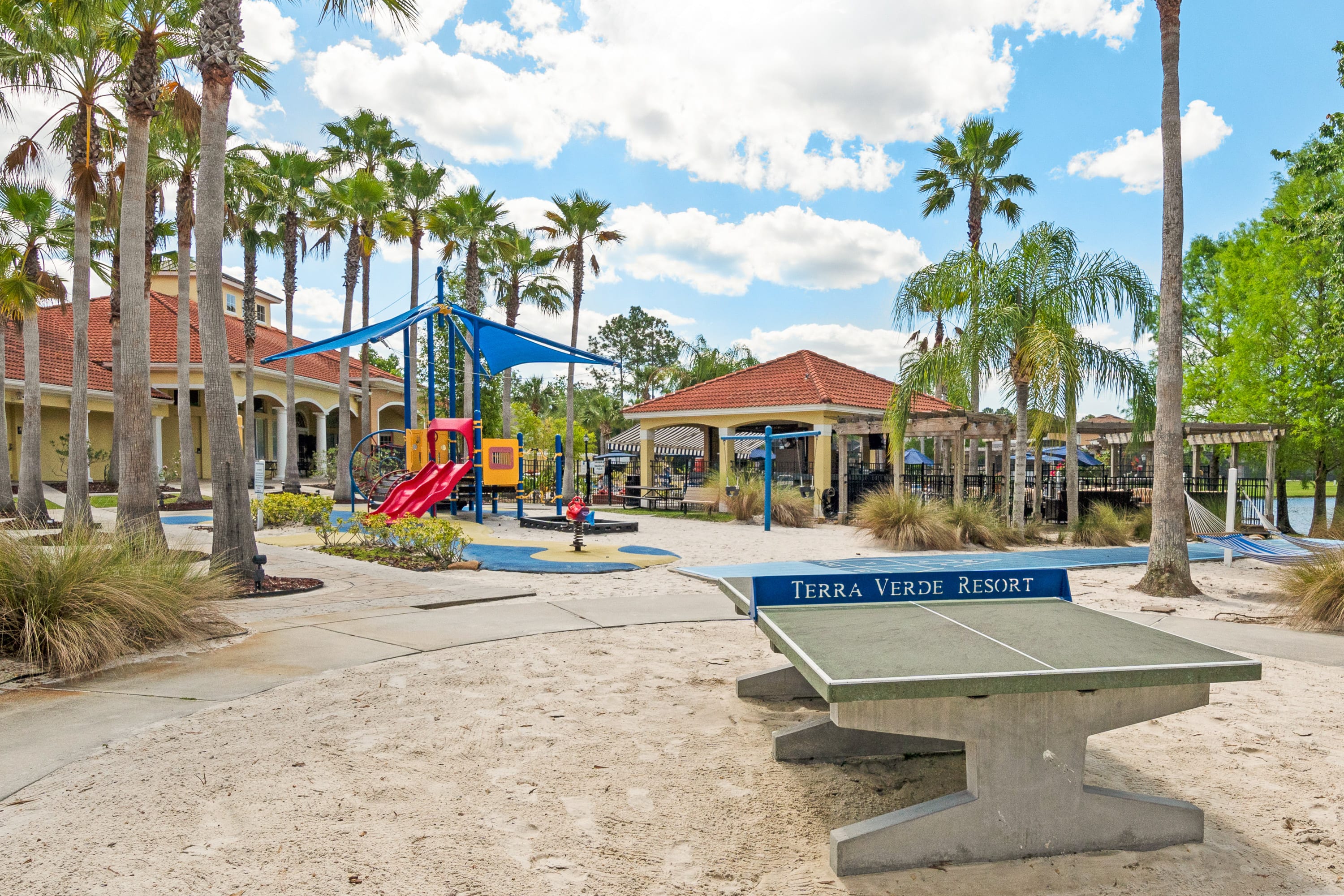 A sandy outdoor recreation area features a colorful children's playground with slides and swings, alongside a concrete ping-pong table with "TERRA VERDE RESORT" written on it. Palm trees and resort buildings are visible under a bright sky.