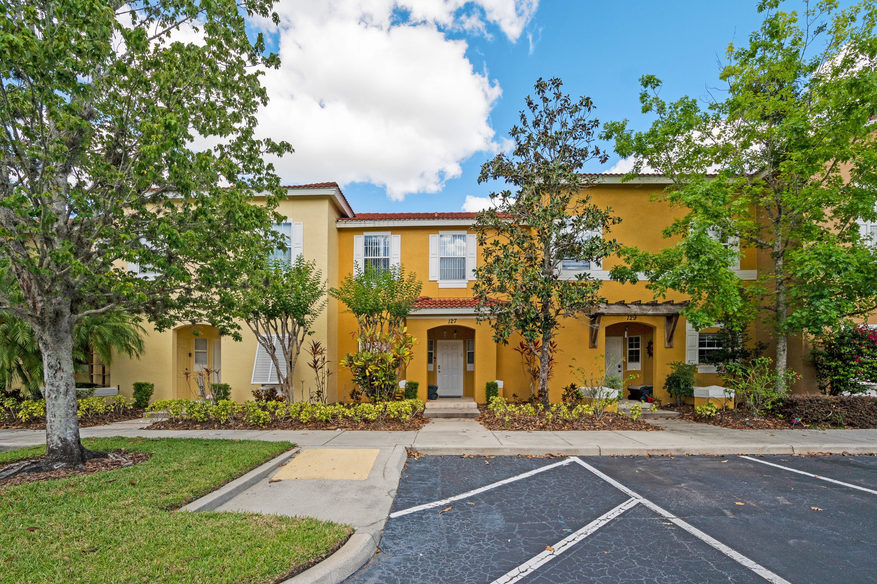 Front exterior view of a two-story townhouse with a warm yellow stucco facade, white window shutters, and a red tile roof. The entrance is framed by lush green landscaping, leading to a paved parking area in the foreground.