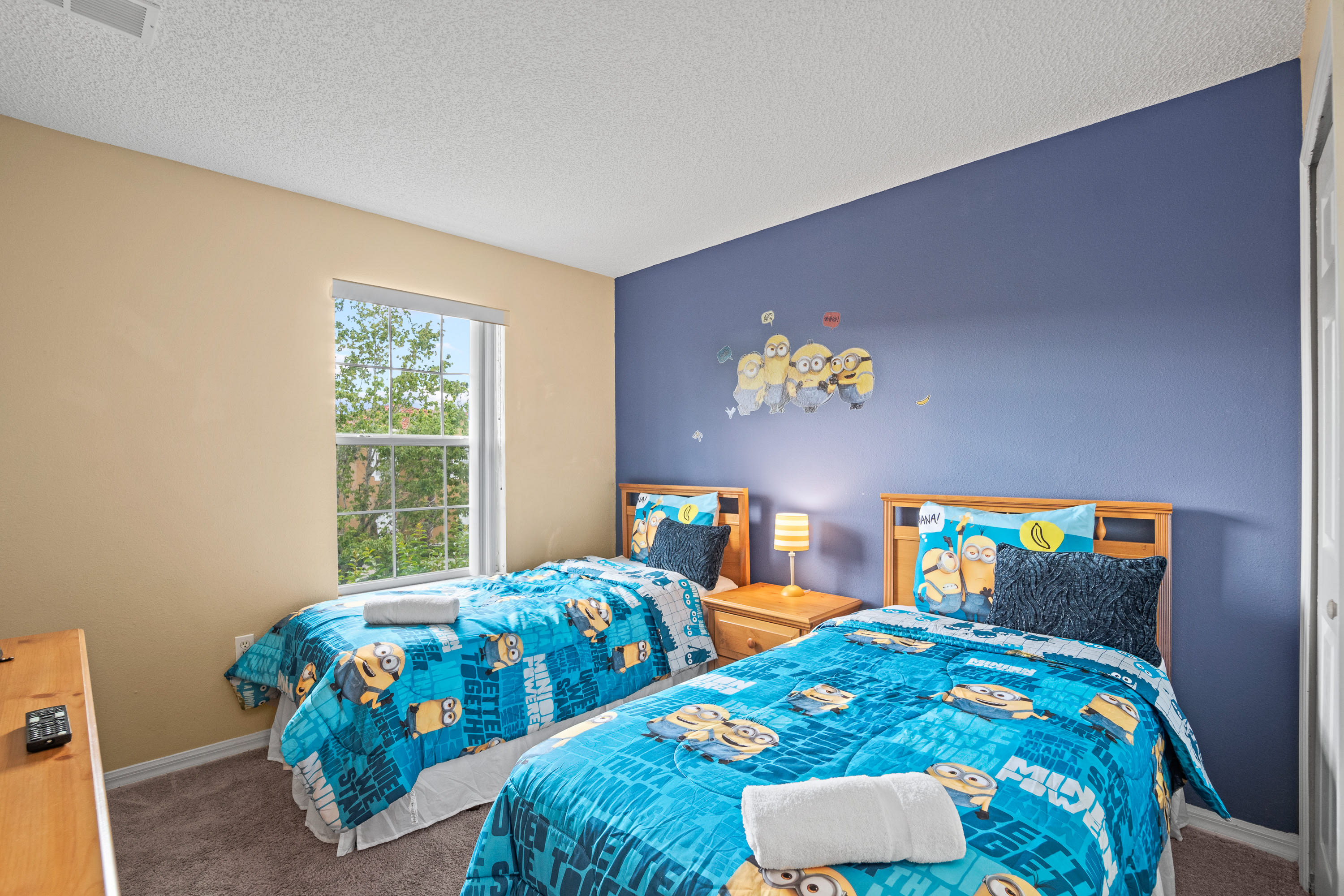 Vibrant kids' bedroom with two twin beds featuring bright blue Minion-themed bedding and pillows, light wood headboards, and a small nightstand with a striped lamp. A large Minion wall decal decorates the blue accent wall.