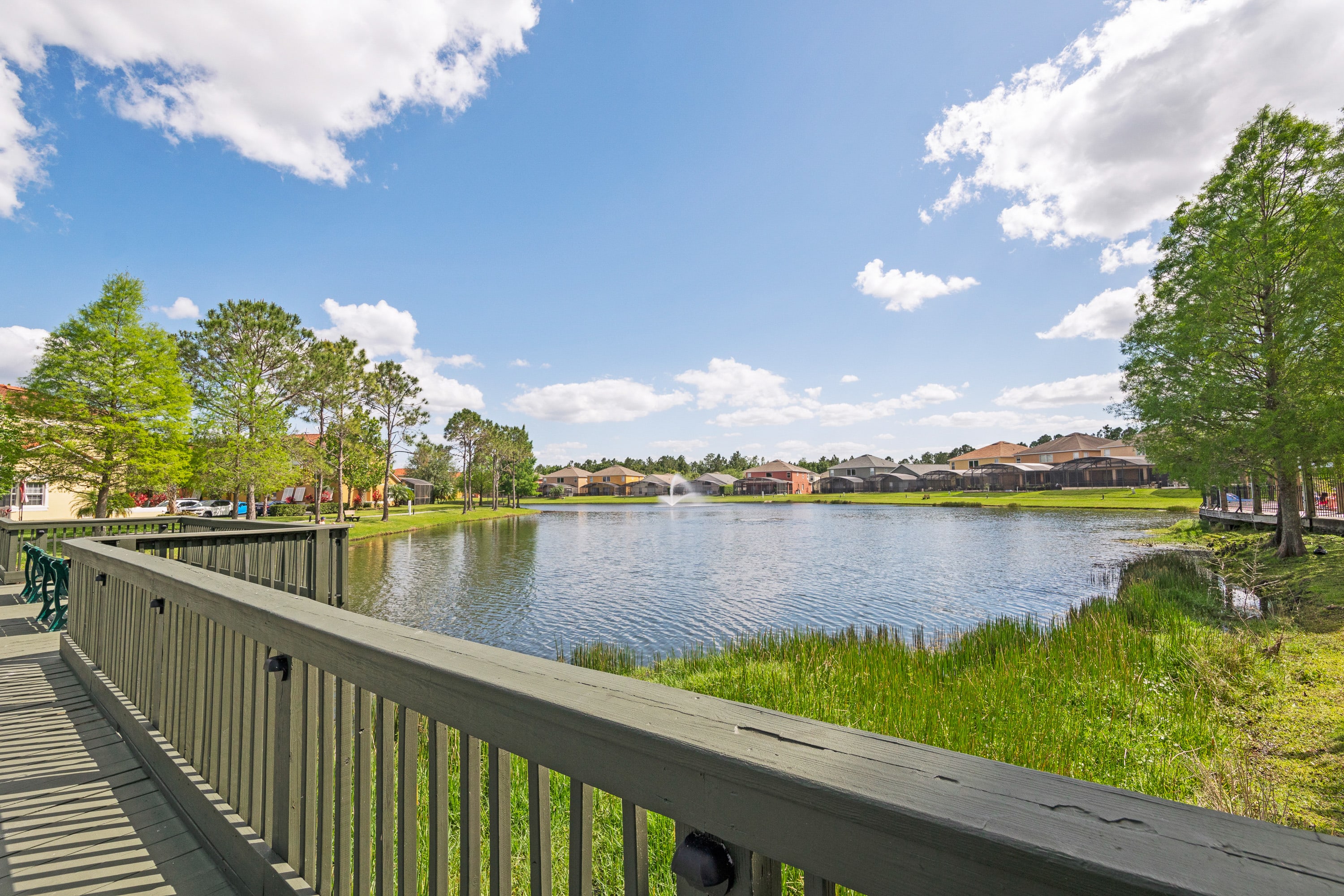 A scenic view overlooks a tranquil lake with a central fountain, bordered by residential homes and lush greenery under a bright blue sky with scattered clouds. A dark green wooden railing is prominent in the foreground.