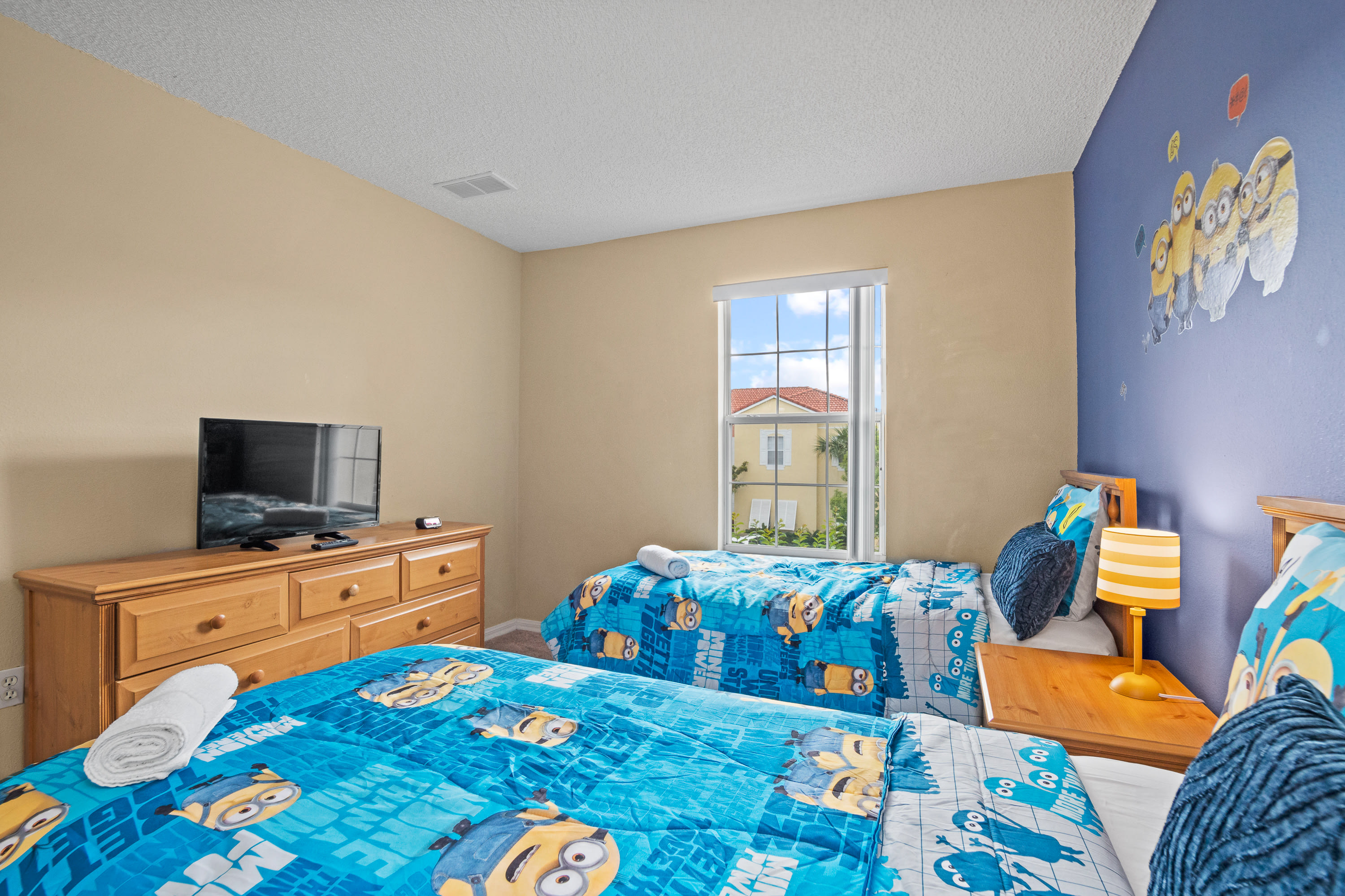 Kids' bedroom showcasing two twin beds with blue Minion bedding, a light wood dresser with a flat-screen TV, and a window overlooking the neighborhood. The room has light yellow walls and a playful blue accent wall.