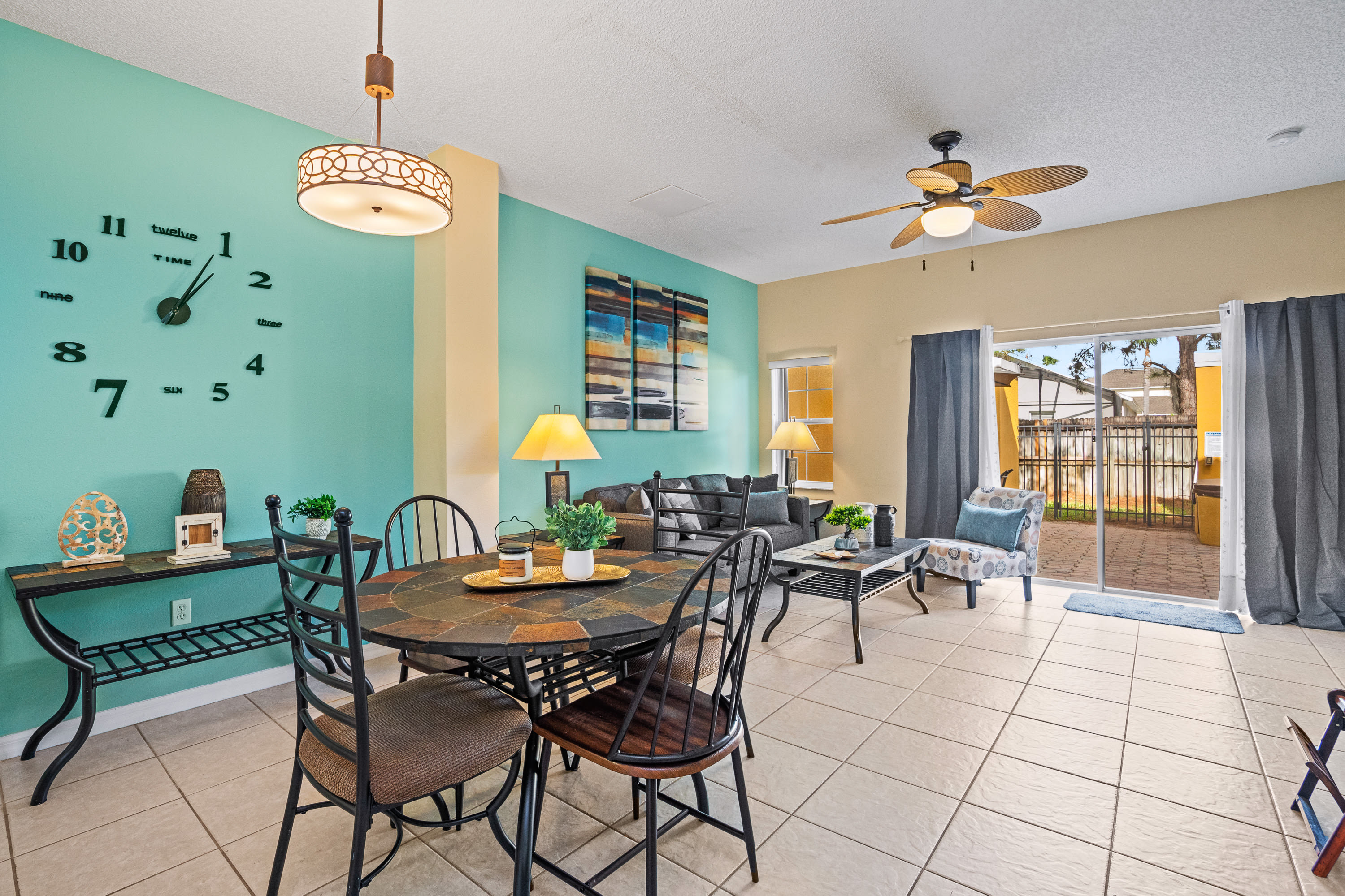 A vibrant dining area with a round, dark-tiled table and four wrought-iron chairs, set against a teal accent wall with a large decorative clock. A living room with a sofa and patio access is visible in the background.