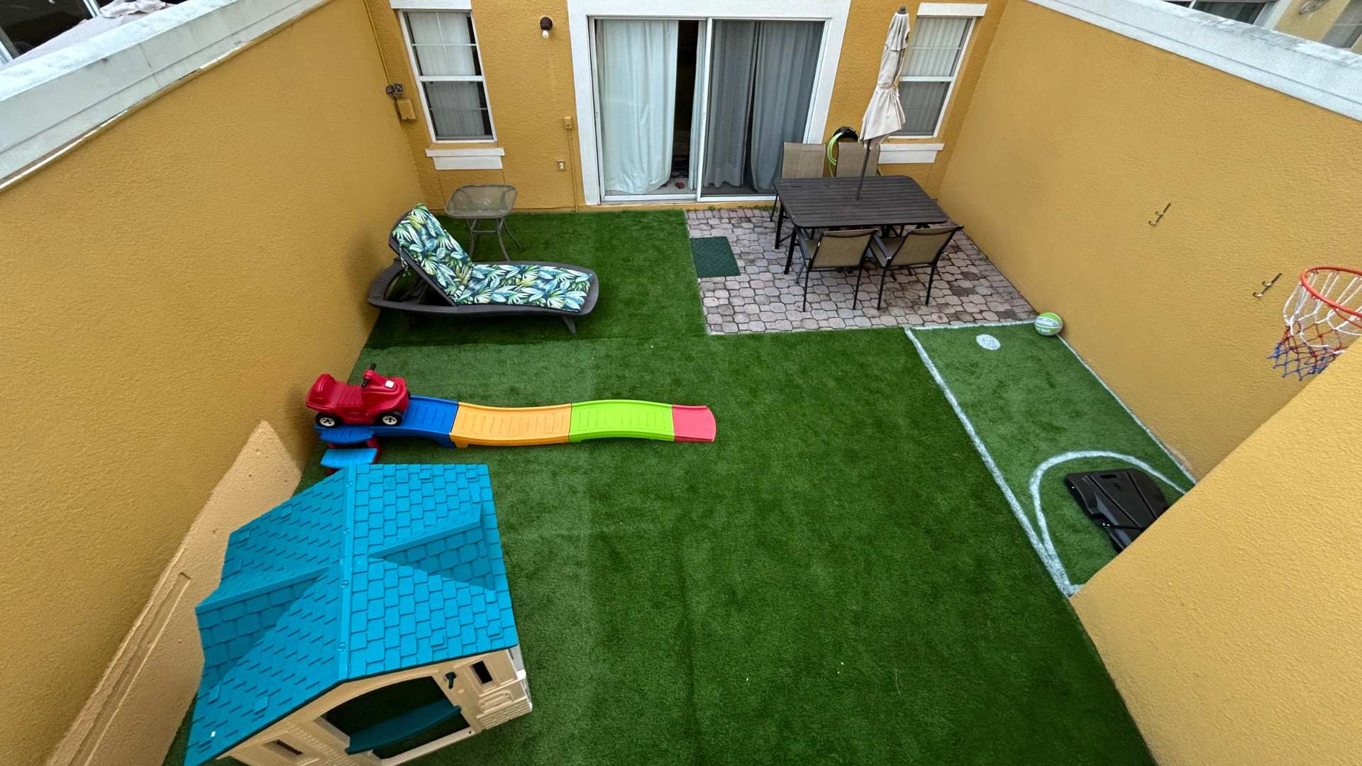 Overhead view of an enclosed outdoor patio with artificial turf, a small playhouse, a slide, and a basketball hoop. A paved area with an outdoor dining table and lounge chair is also visible, leading to a sliding glass door.